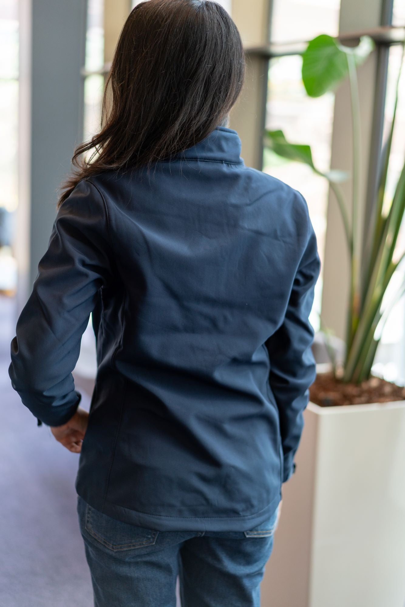 Back view of a female model with her hair down wearing a navy softshell jacket from the RMIT Store. The jacket features a clean, tailored fit with subtle seam detailing and a high collar, shown against an office background.