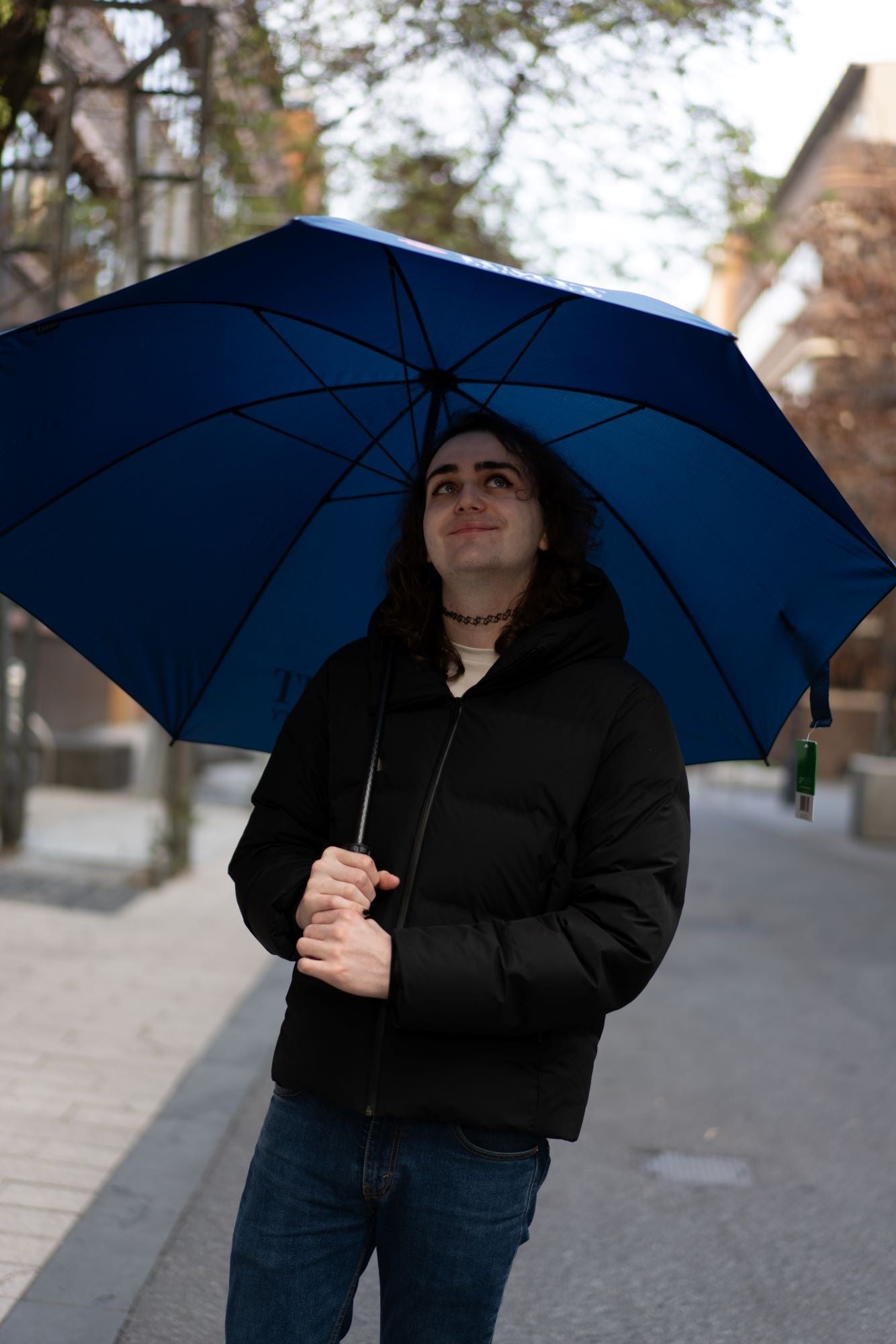 Close-up view of a model wearing blue denim jeans and a black jacket, holding a navy umbrella with a black handle. The model is looking up under the umbrella, smiling. The navy blue canopy of the umbrella is visible, and the model’s grip on the handle is clear. The background is blurred, focusing on the model and the umbrella. 
