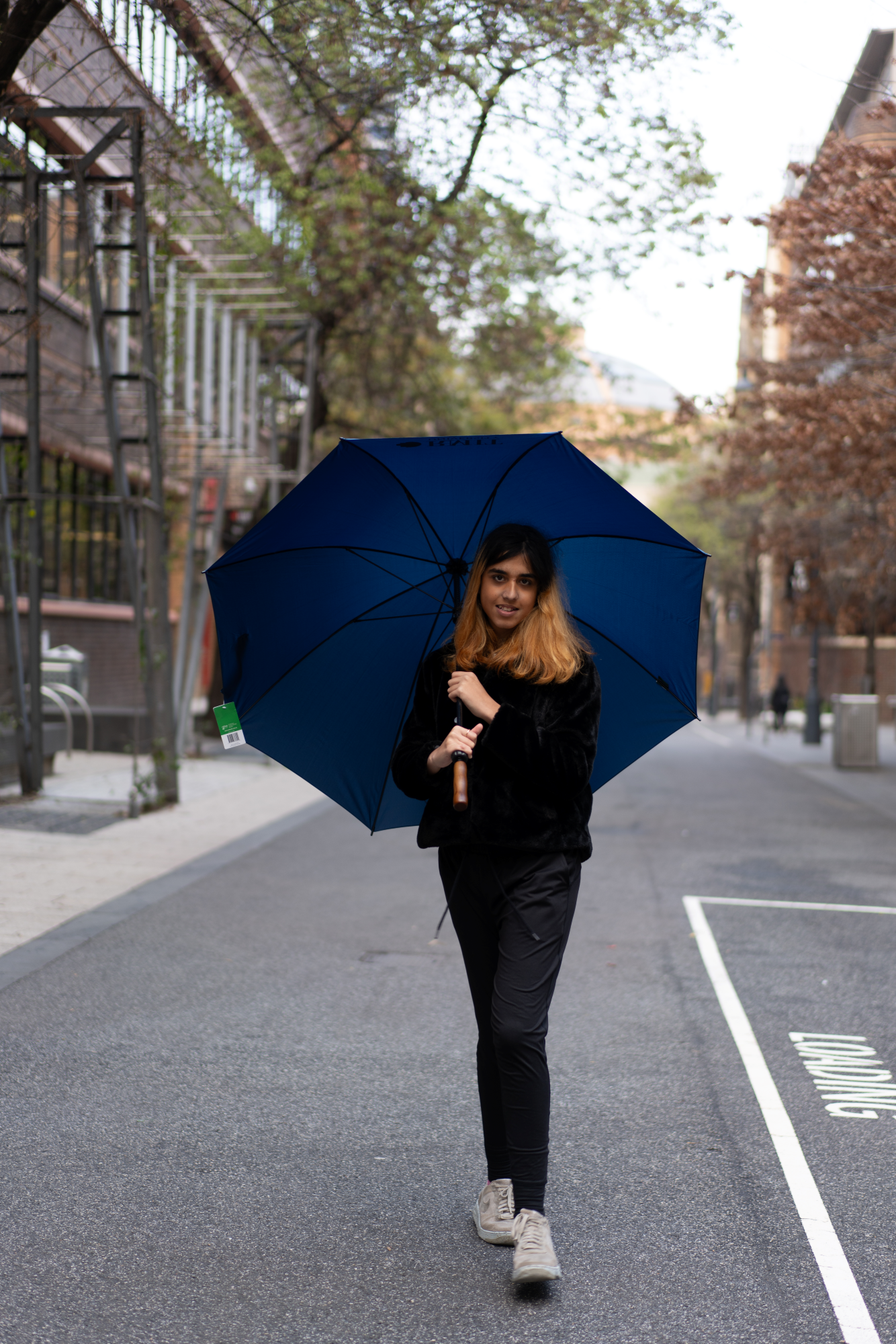 Navy umbrella held by a model wearing all black clothing, walking on campus. The model is gripping the black handle of the umbrella, which is open, with the deep navy blue canopy visible. The background shows a university campus setting, with trees and buildings in the distance.