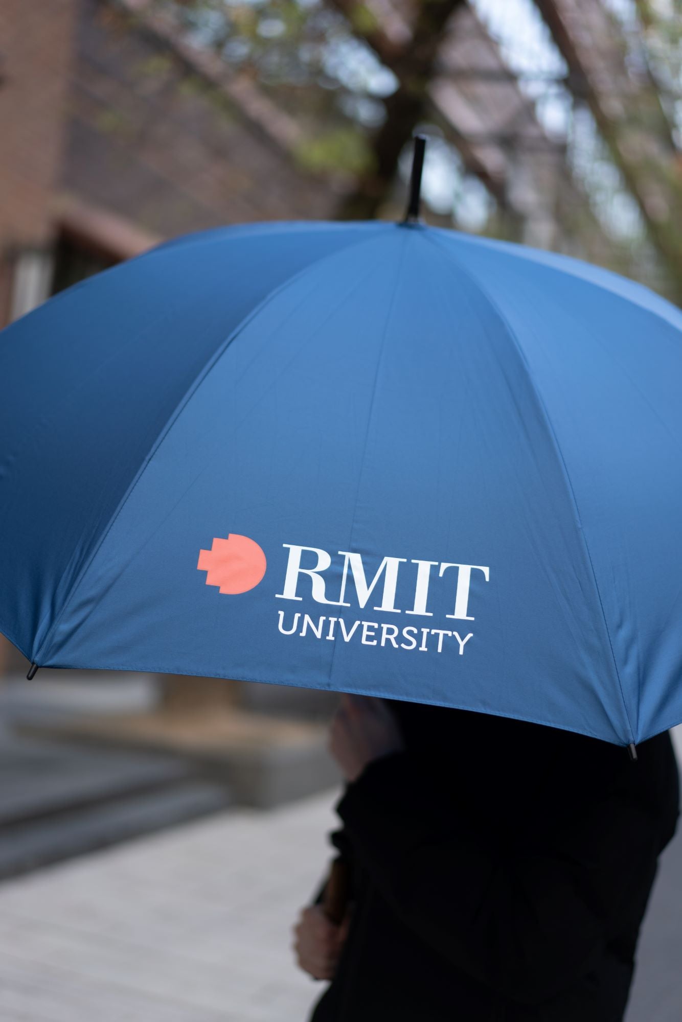 Close-up view of one side of a navy umbrella, with the RMIT University logo in white and red visible on the fabric. The umbrella's fabric is smooth, showing its navy blue colour, with the logo prominently displayed. The focus is on the logo, which is located on the side of the umbrella. The background is a blurred campus setting, providing a clean view of the umbrella.