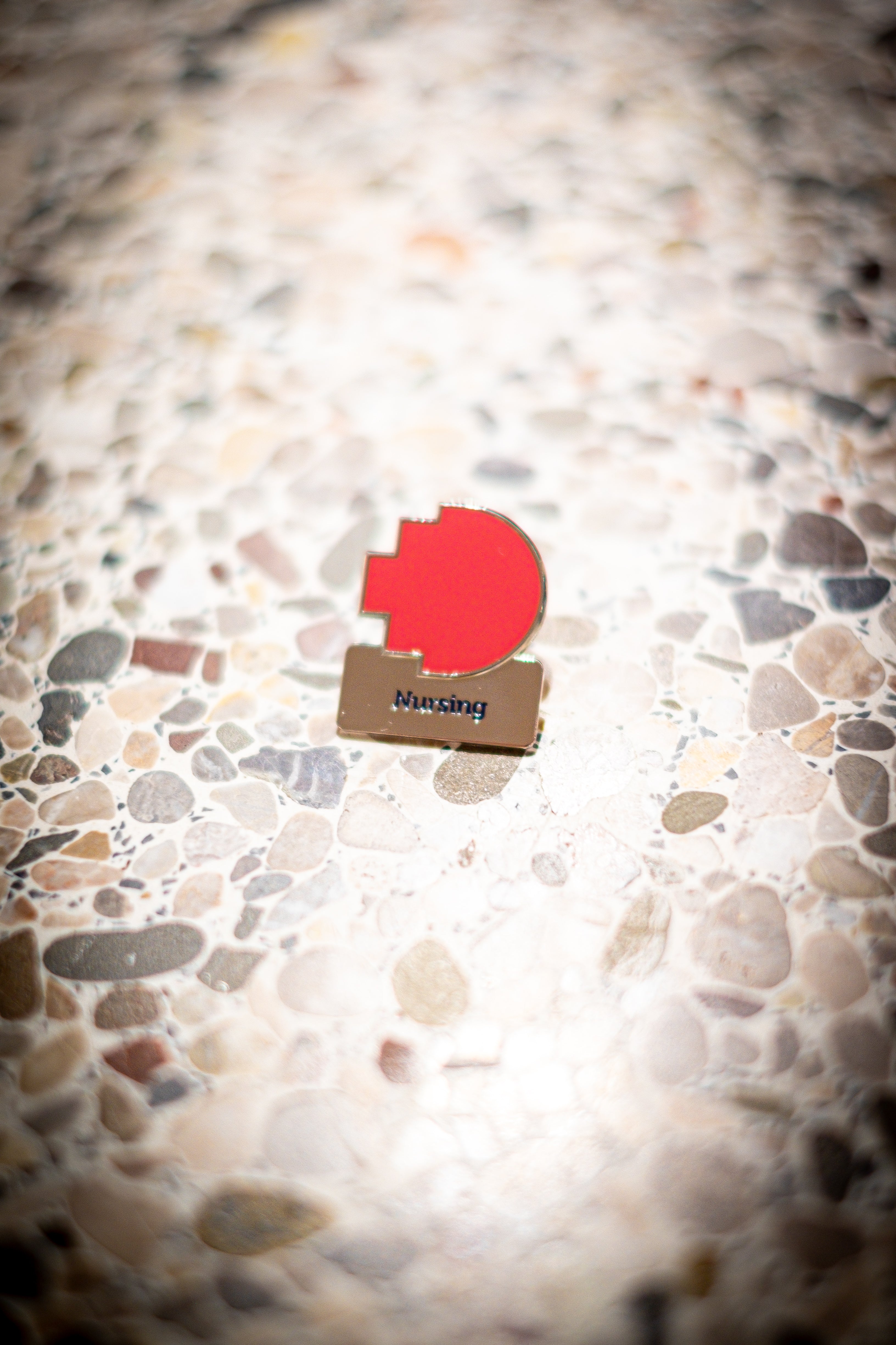 Nursing discipline pin featuring a red pixelated logo above a silver square with 'Nursing' text, resting on a light marble background, available at the RMIT Store.