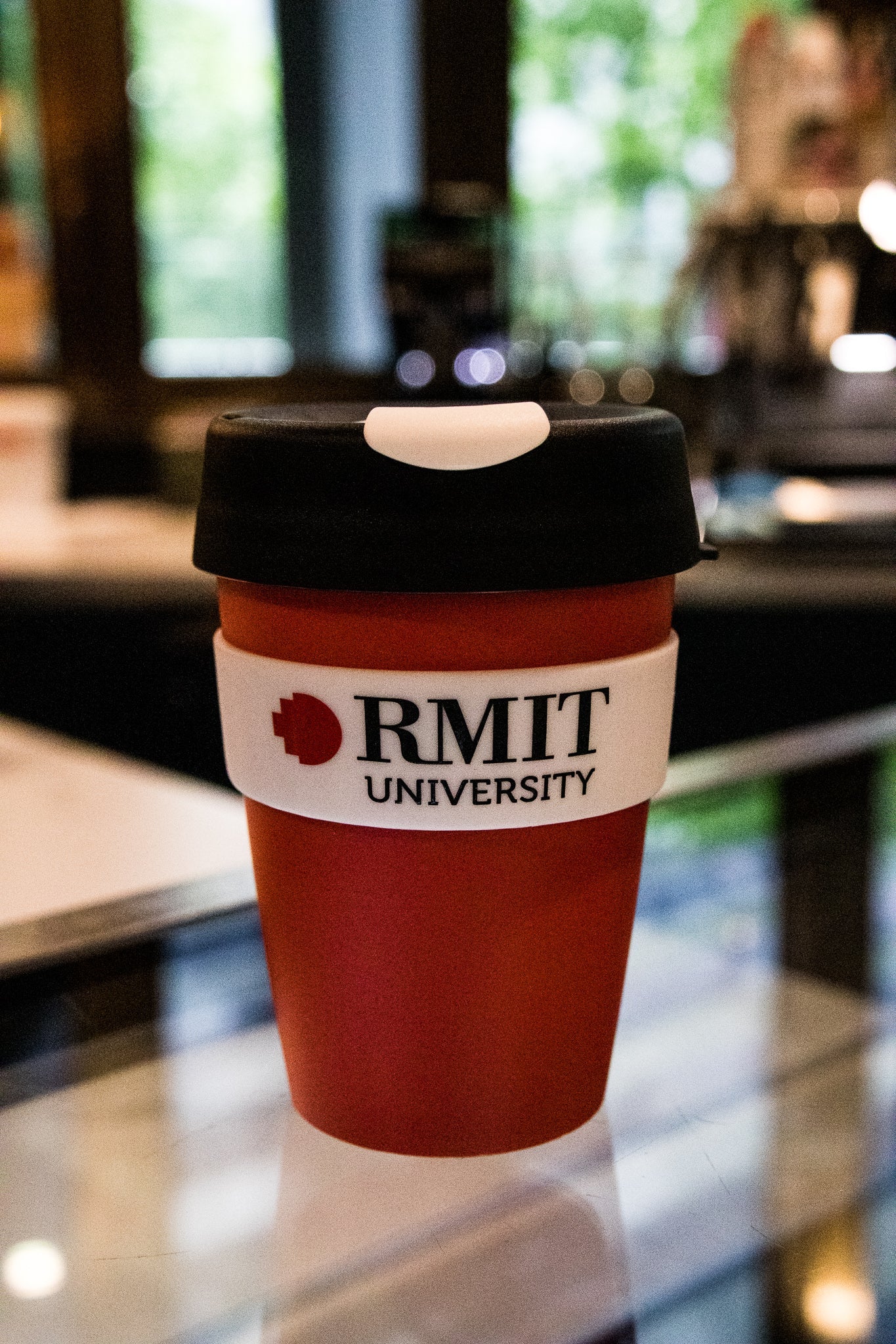 Original RMIT KeepCup with red body and black lid, adorned with white branding, situated on a café countertop with blurred background.