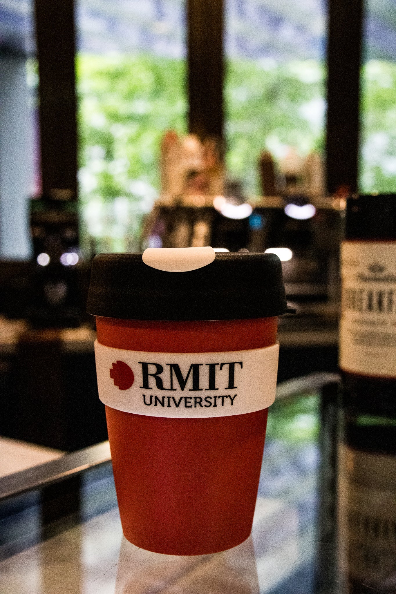 Front view of the Original RMIT KeepCup, red with RMIT University branding, placed on a glass counter with a coffee machine in the background.