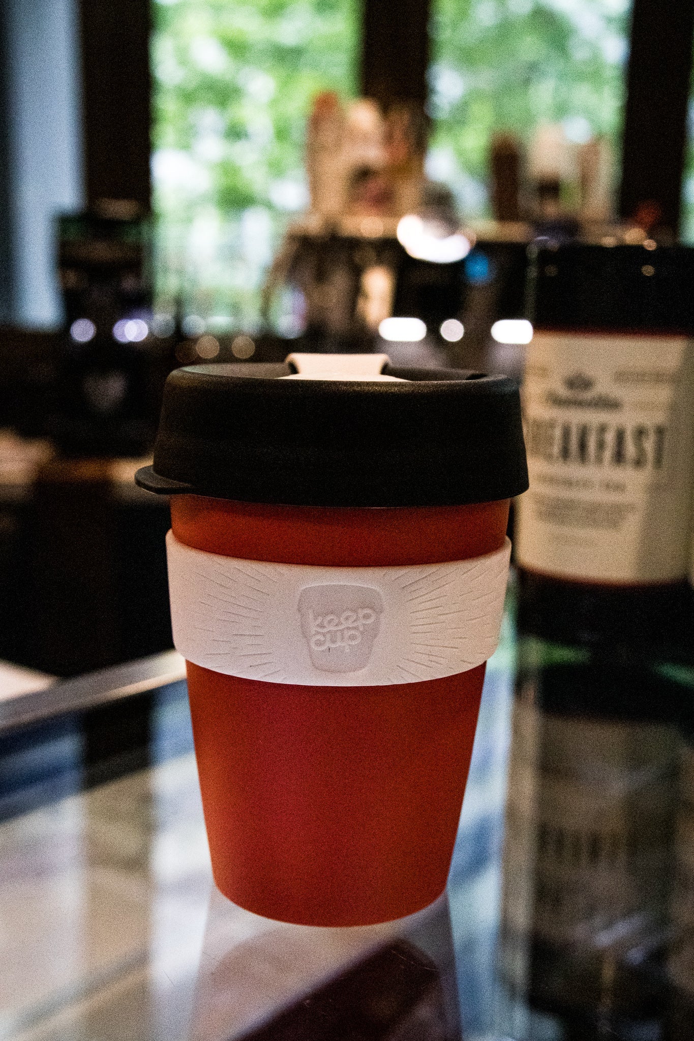 Side view of the Original RMIT KeepCup featuring the KeepCup logo on its white band. Red cup with black lid on a glass surface in a café environment.