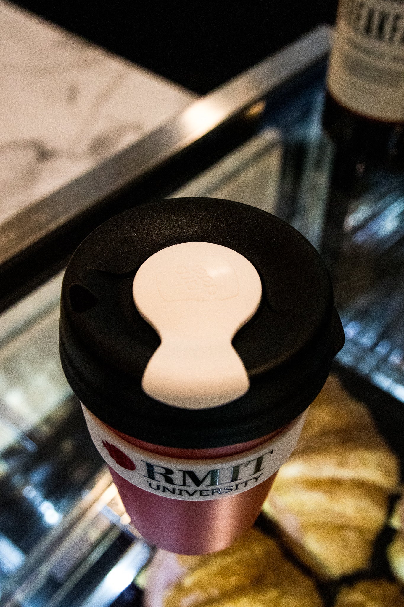 Top view of the Original RMIT KeepCup with a black lid and white seal, showcasing the RMIT University branding. The cup rests on a reflective surface in a café setting.