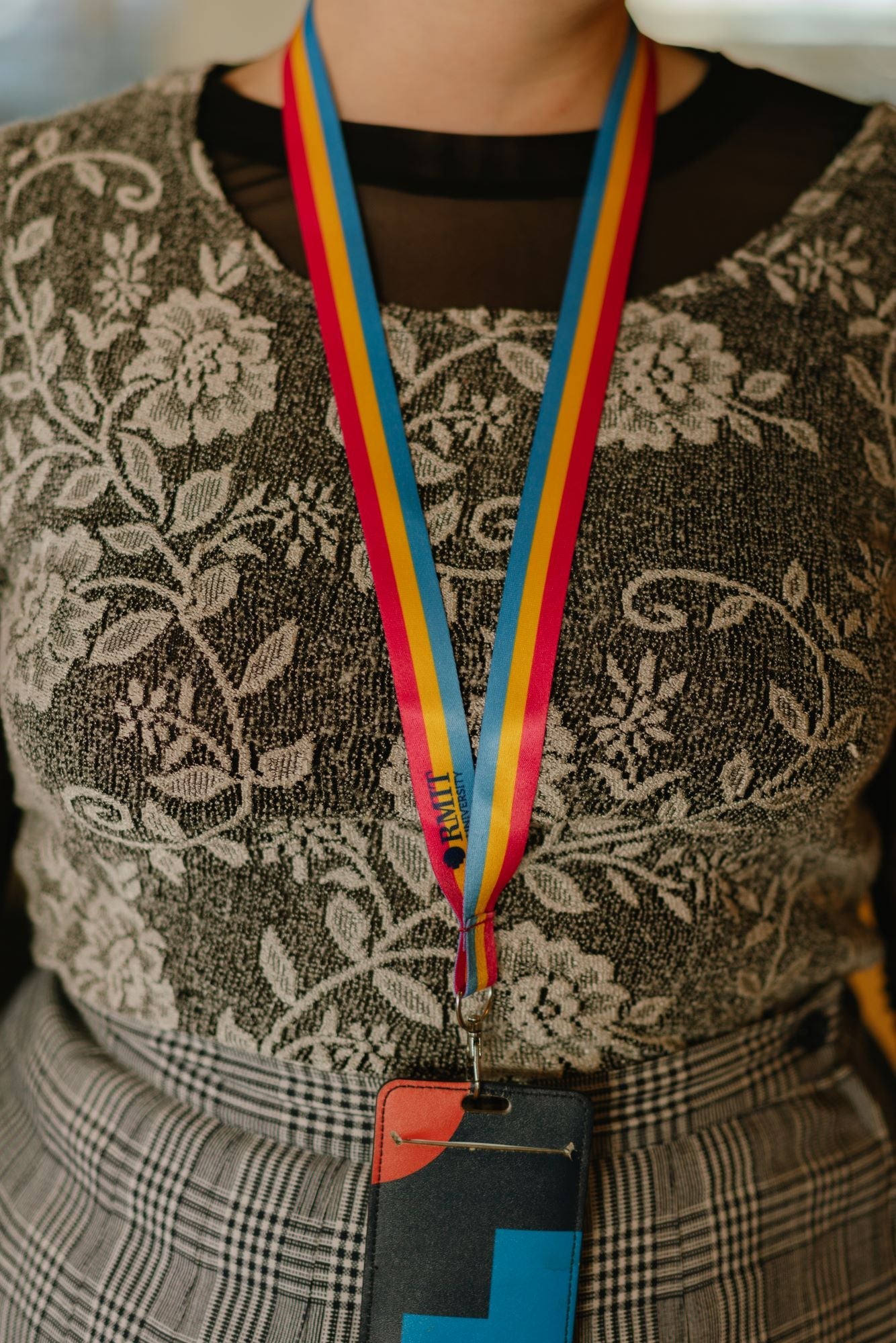 Close-up of a model wearing a Pansexual Pride lanyard from the RMIT Store, featuring horizontal stripes in pink, yellow, and blue with a subtle engraved RMIT logo near the silver metal clip. The lanyard is attached to a card holder. The model wears a brown top.