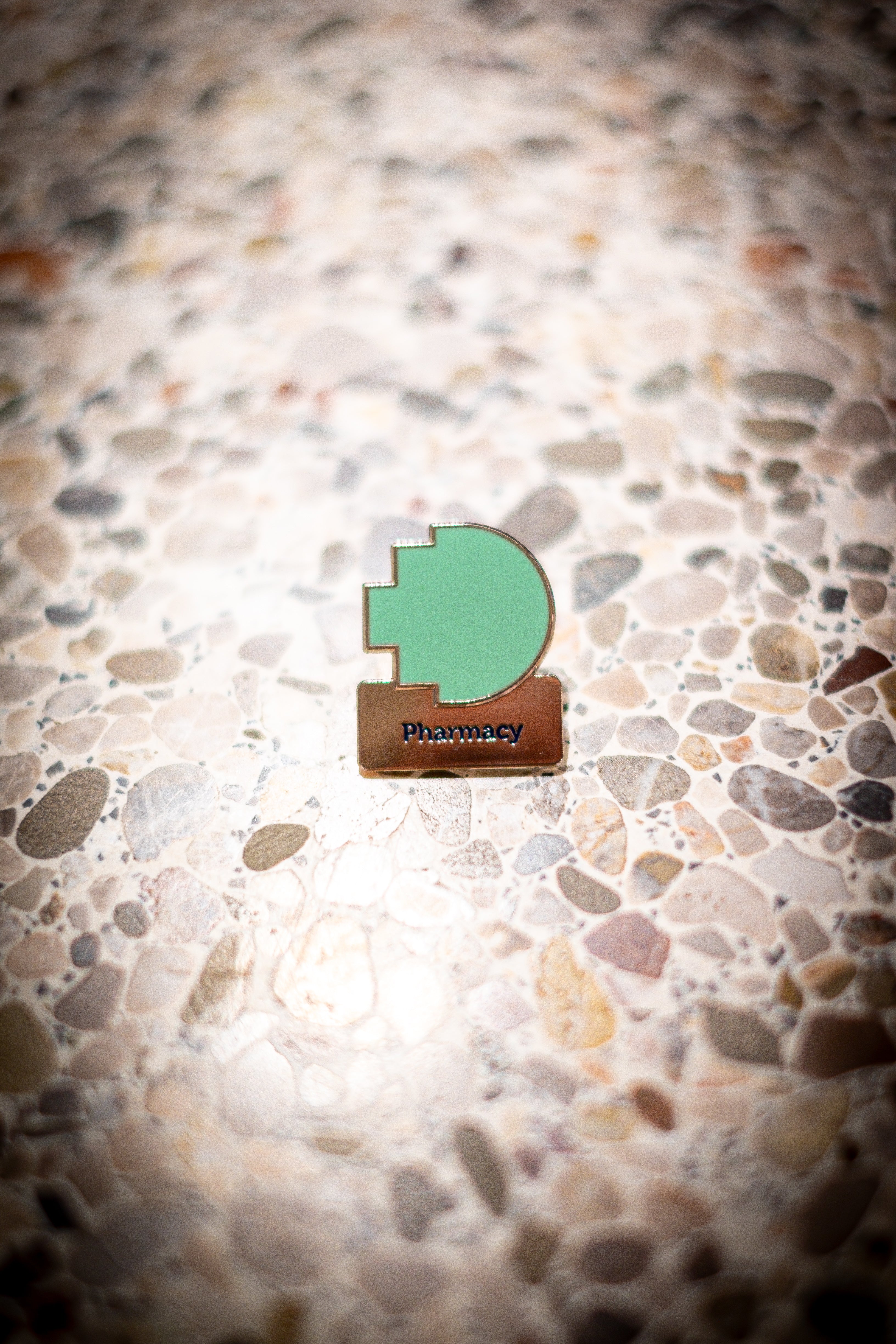 Front view of the Pharmacy Discipline pin on a light marble background, featuring the RMIT pixel logo in green above a silver square with the word "Pharmacy". Available at the RMIT Store.