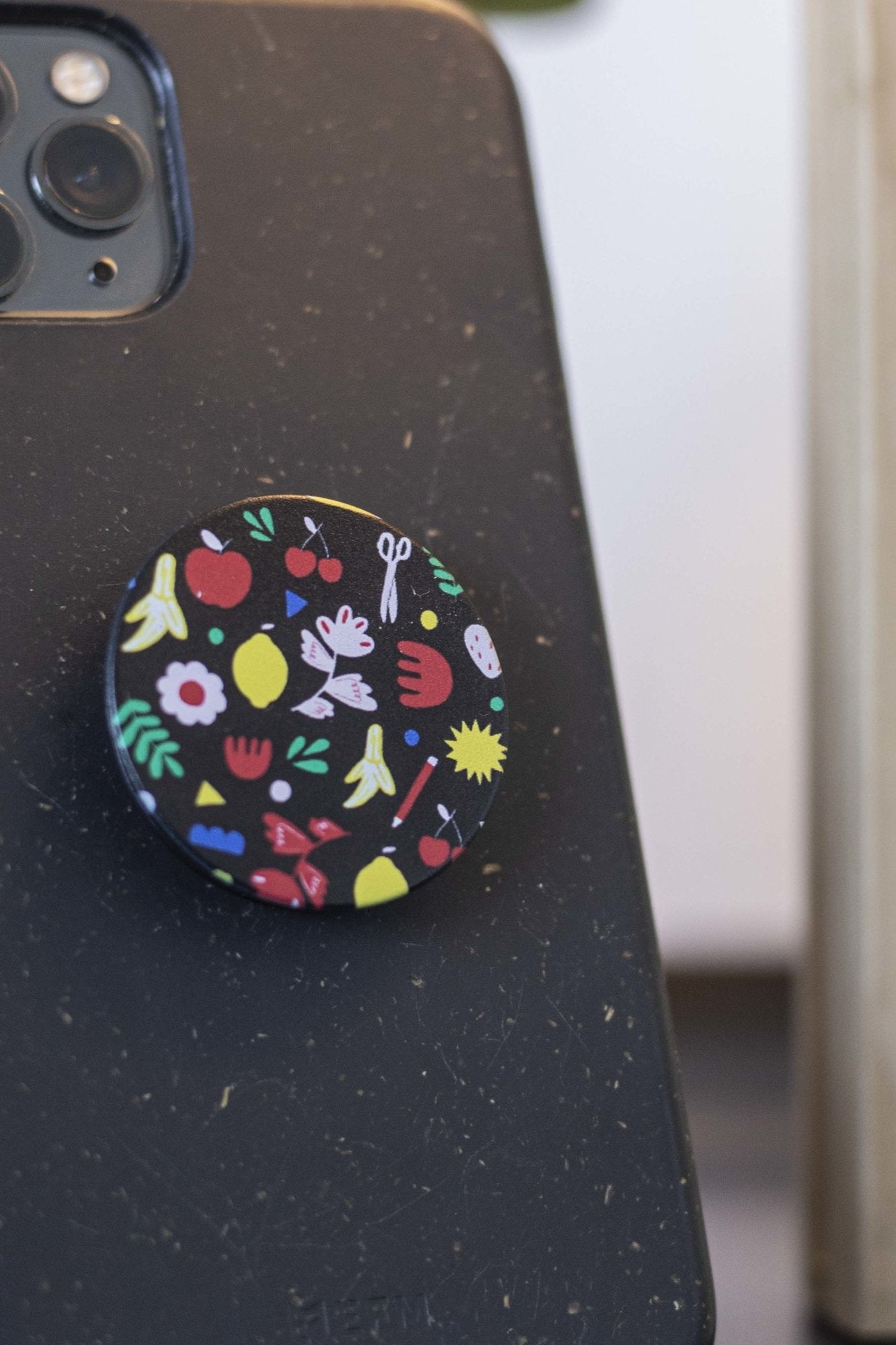 Front close-up of an RMIT Store Isabella Manakis Designed Phone Grip with a black circular base and a colourful abstract patterns, attached to the back of a black smartphone.