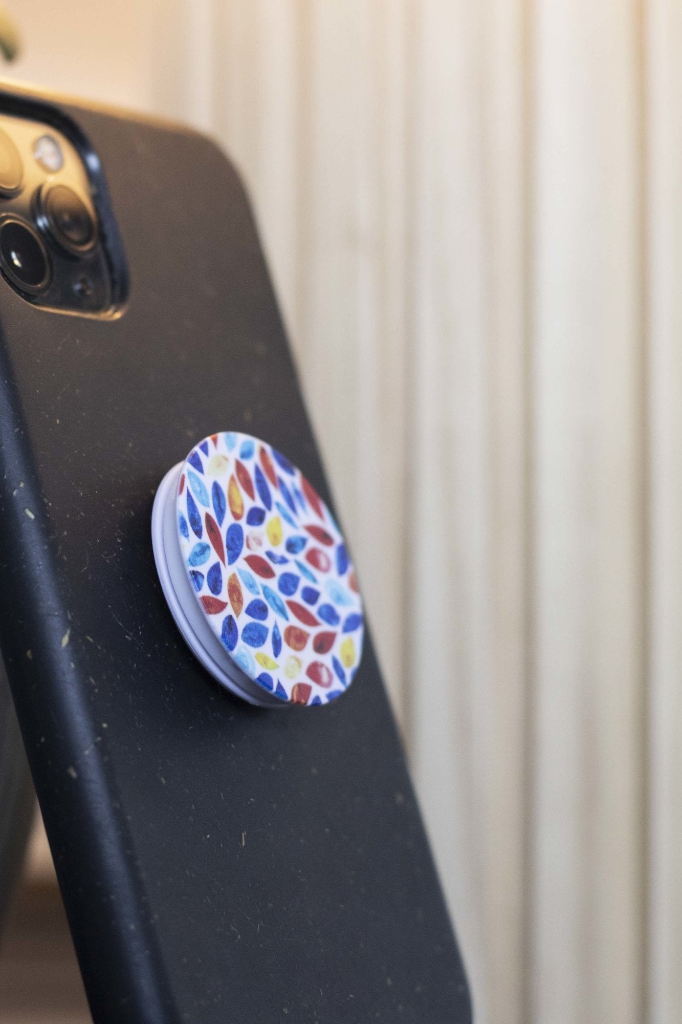Front close-up of two RMIT Store Leaves of Change Phone Grips with a white circular base and red, blue, and yellow leaf designs, attached to the back of a black smartphone.