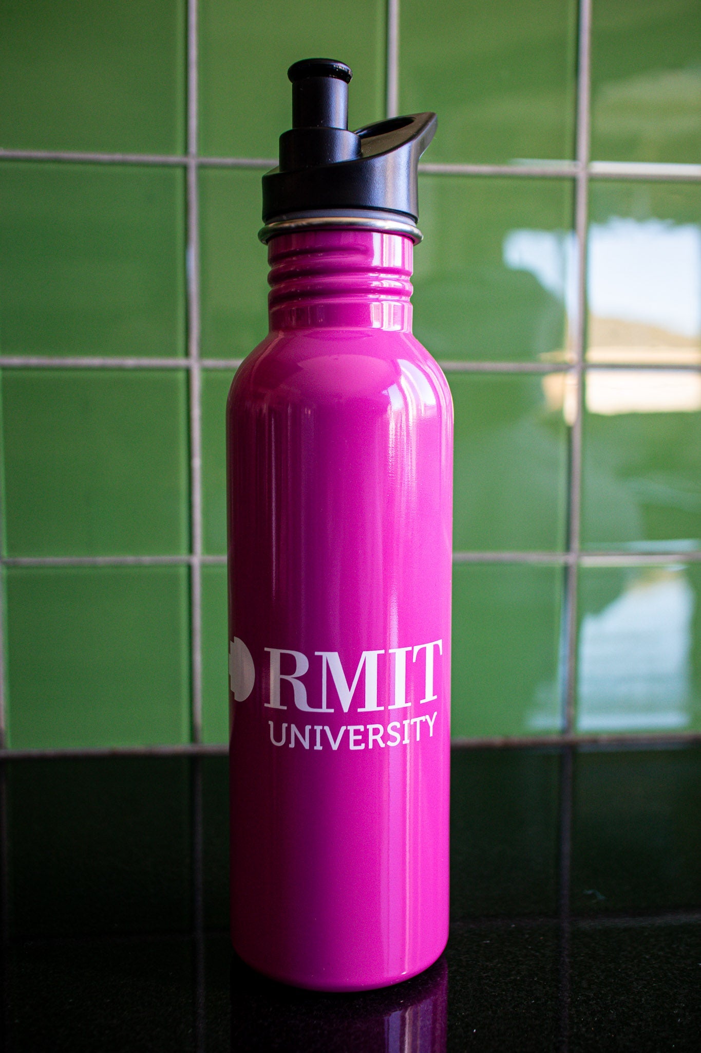 Full view of the pink RMIT bottle with a white RMIT University logo centered on the body, black cap securely on the bottle, placed on a black tabletop with a green background. Available at the RMIT Store.