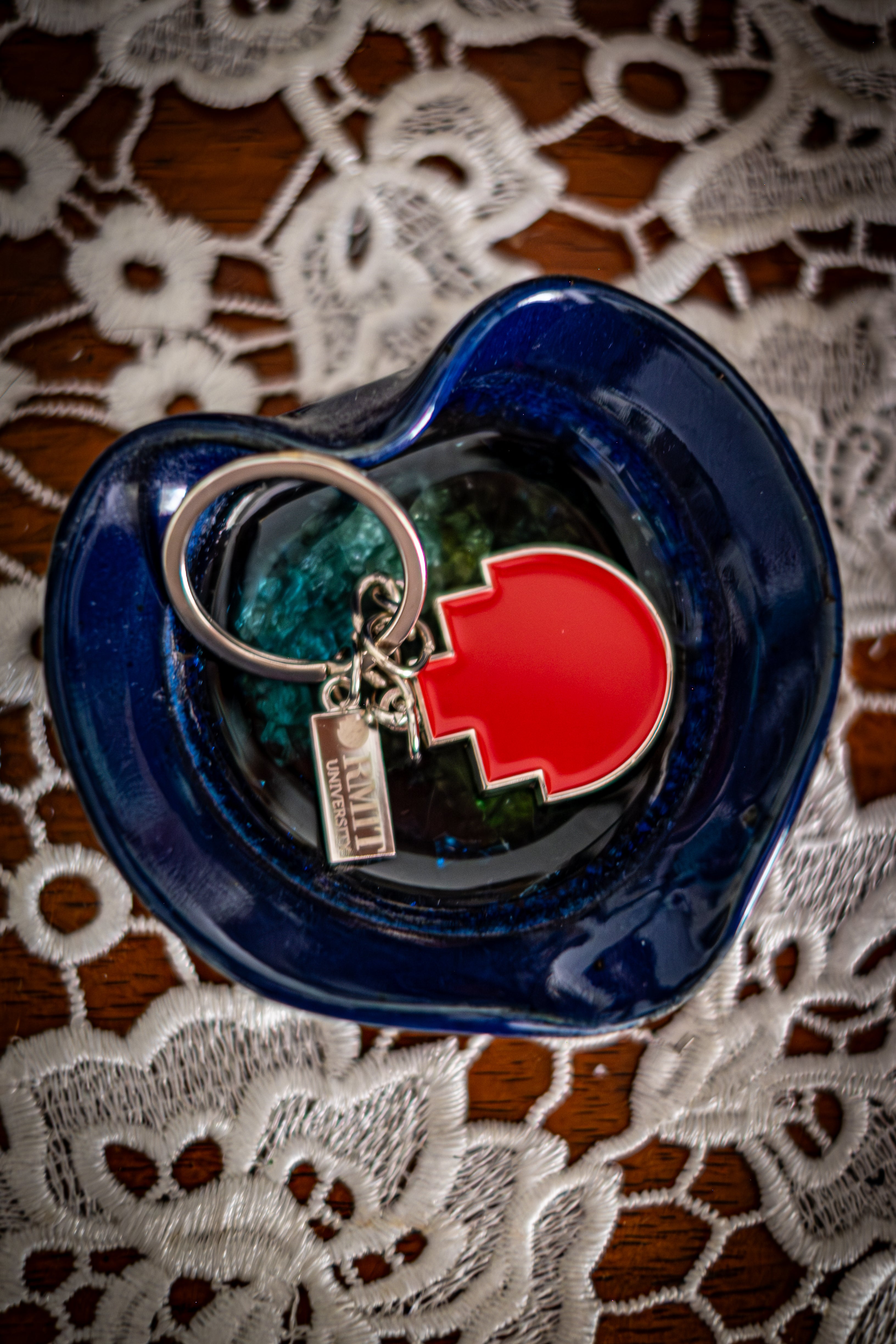 RMIT Pixel Keyring featuring a red pixel-shaped charm with a small silver tag engraved with “RMIT University,” displayed inside a glossy dark blue ceramic dish on a white lace-covered wooden surface. Available at the RMIT Store.
