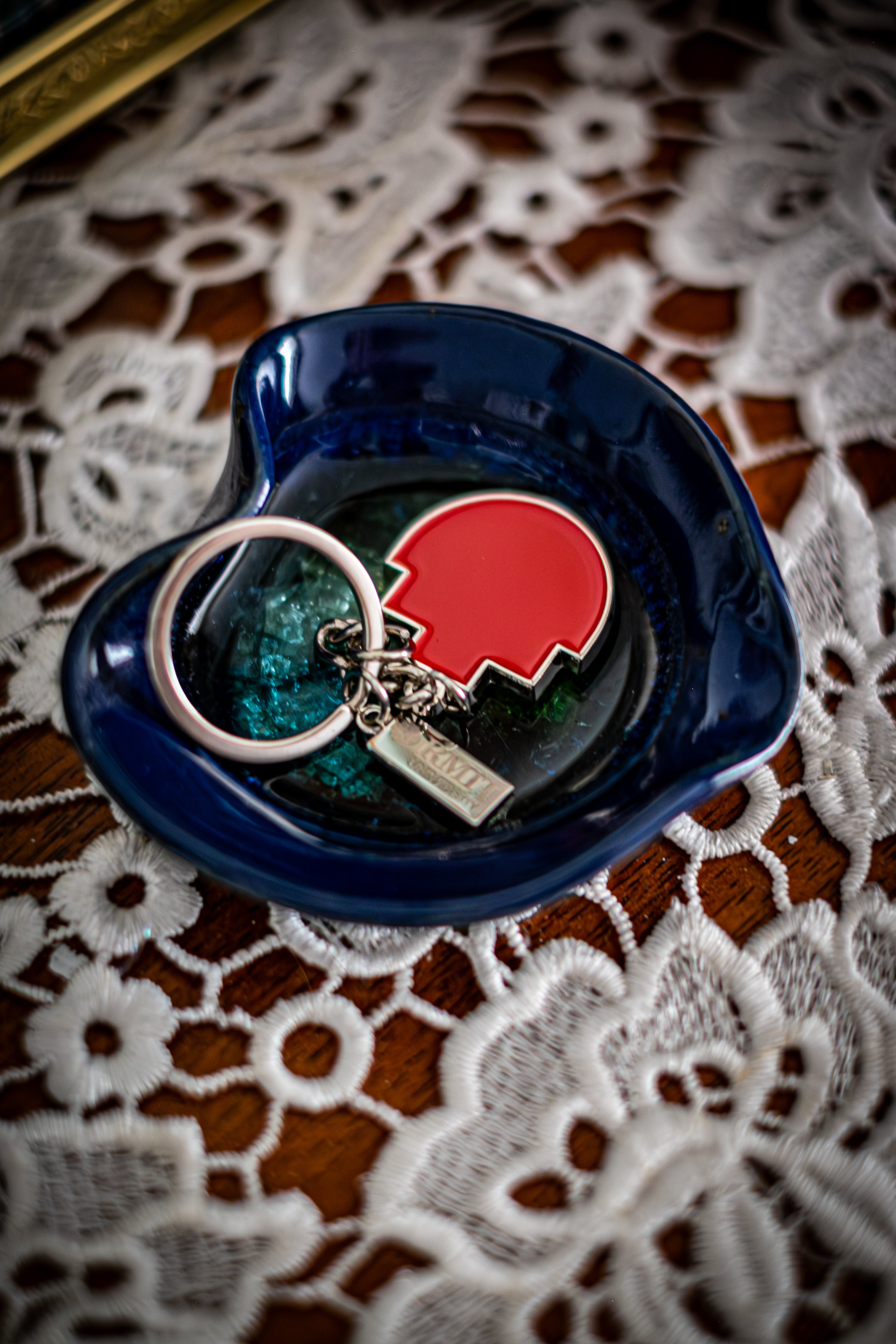 RMIT Pixel Keyring with red enamel pixel-shaped charm and small silver tag engraved with “RMIT University,” displayed flat inside a glossy dark blue ceramic dish on a lace-covered wooden surface. Available at the RMIT Store.