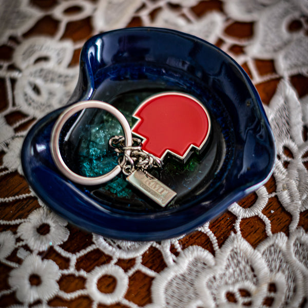 RMIT Pixel Keyring with red enamel pixel-shaped charm and small silver tag engraved with “RMIT University,” displayed flat inside a glossy dark blue ceramic dish on a lace-covered wooden surface. Available at the RMIT Store.