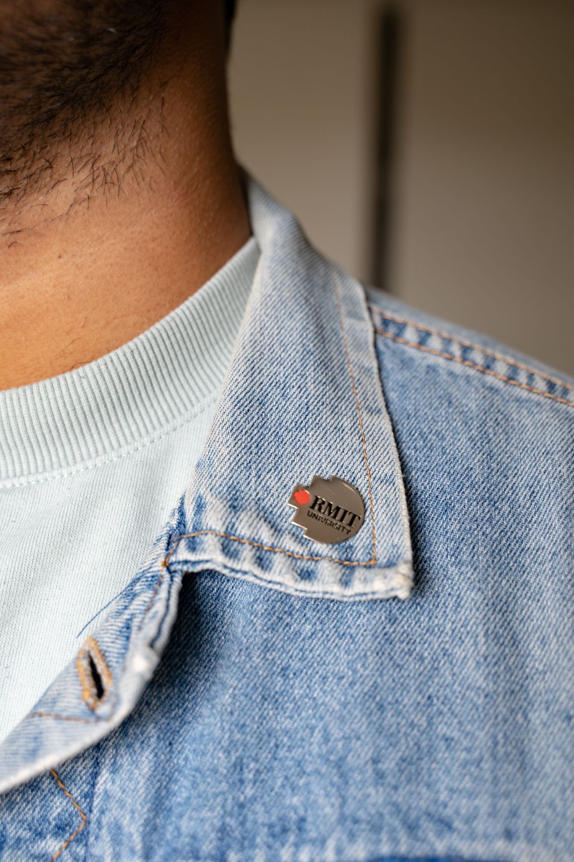 Model showcasing the RMIT Pixel Lapel Pin on a denim jacket collar. The pin displays the RMIT logo against a red and grey backdrop, offered by the RMIT Store for a sleek accessory.
