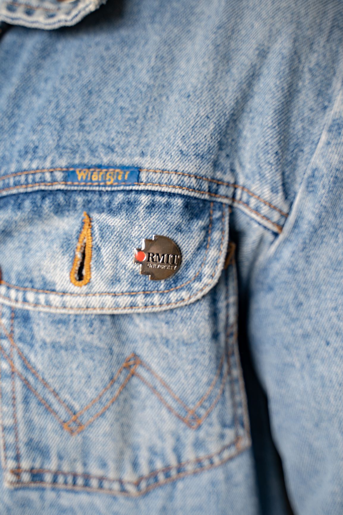 Model wearing the RMIT Pixel Lapel Pin on a blue denim jacket pocket. The pin features the RMIT logo with a red and grey design, highlighting brand loyalty from the RMIT Store.