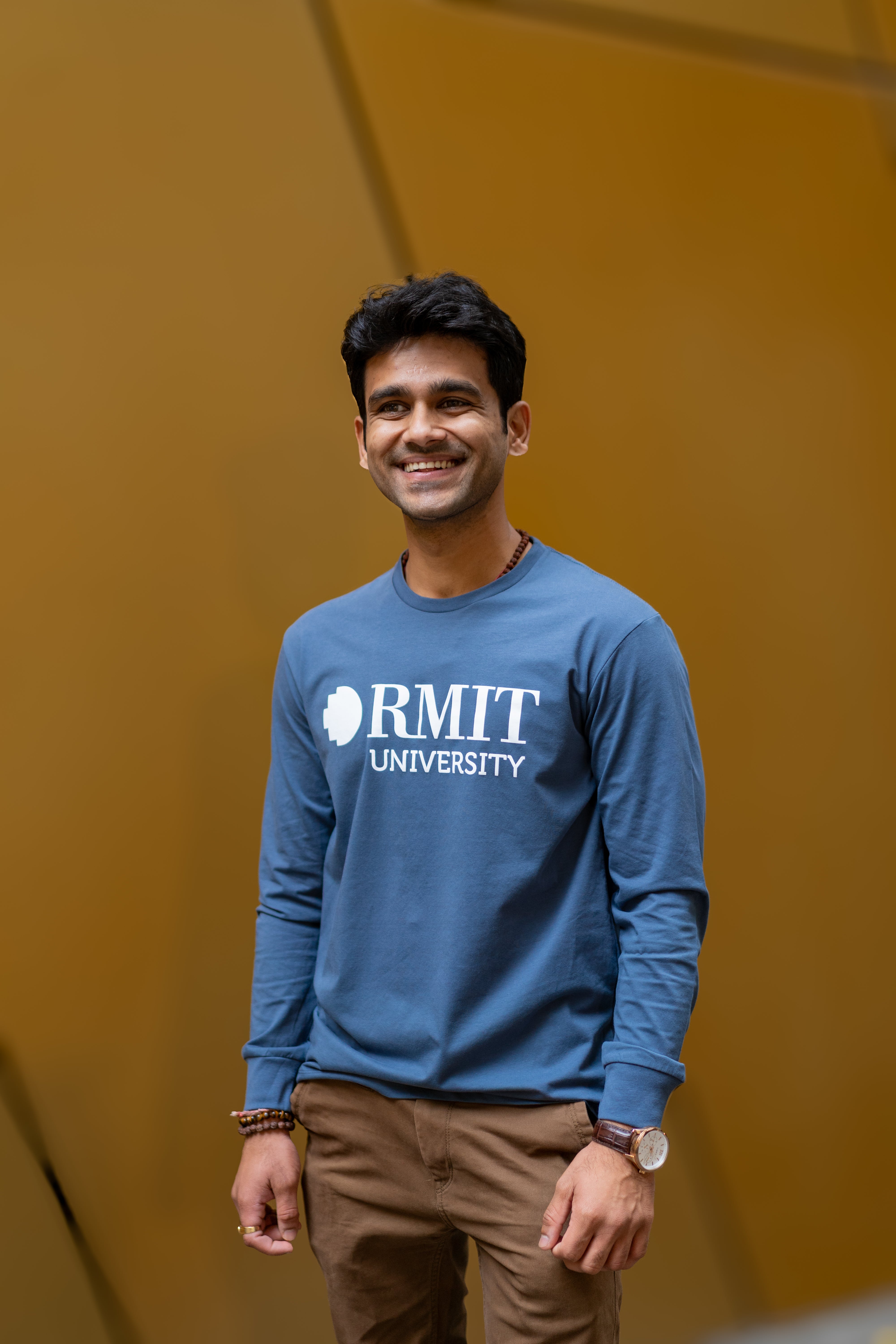 Male model dressed in RMIT Plenty Long Sleeve Tee in slate blue, complemented by light brown trousers and a wooden bracelet and watch.