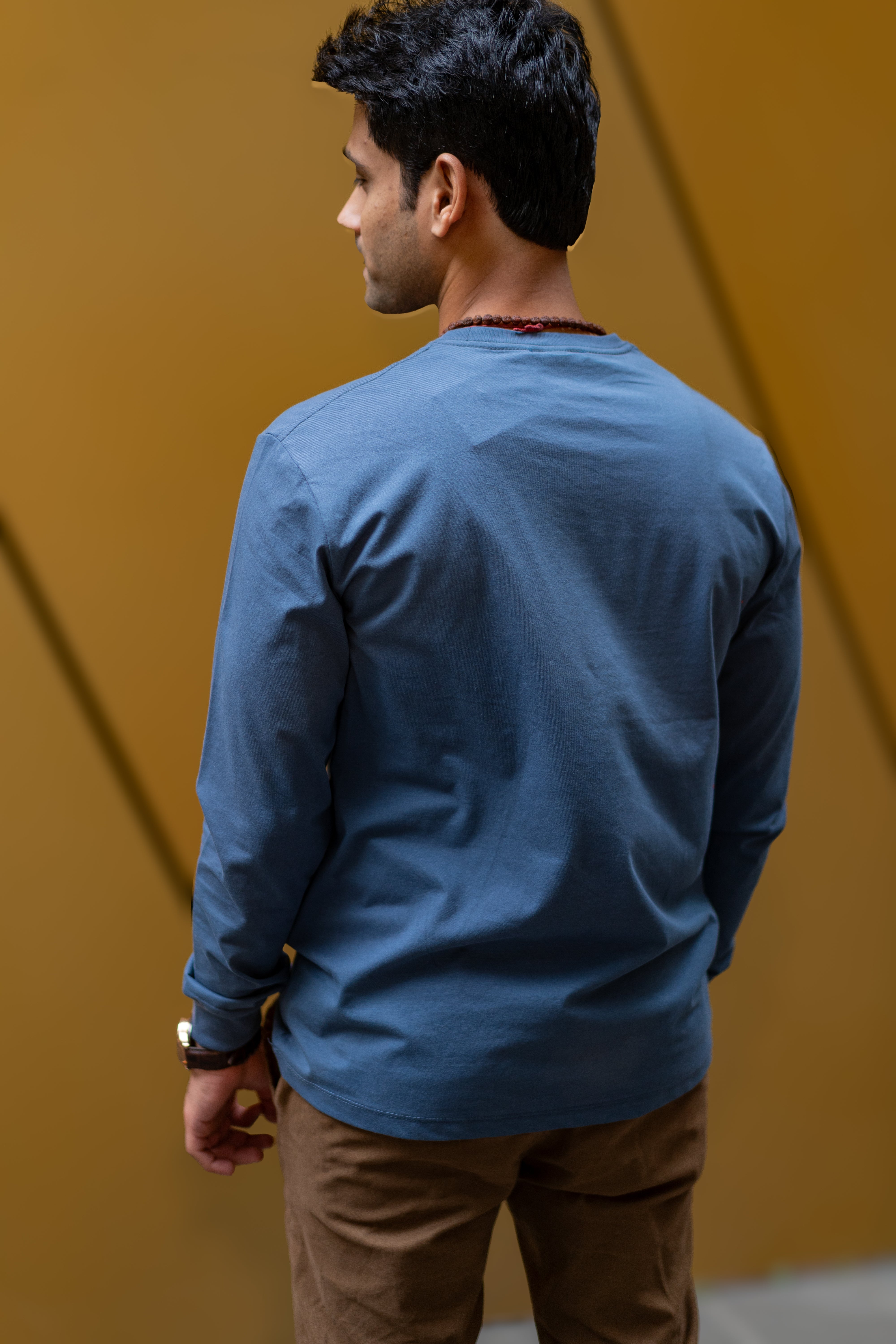 Rear view of a male model in RMIT Plenty Long Sleeve Tee in slate blue, paired with light brown trousers, highlighting the tee's seamless design.
