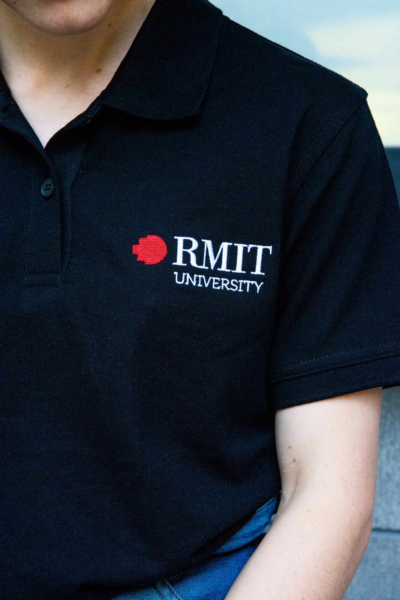 Close-up of the black RMIT Polo, highlighting the RMIT University logo embroidered in red and white on the chest. The fabric texture and stitching detail showcase quality craftsmanship, available at the RMIT Store.