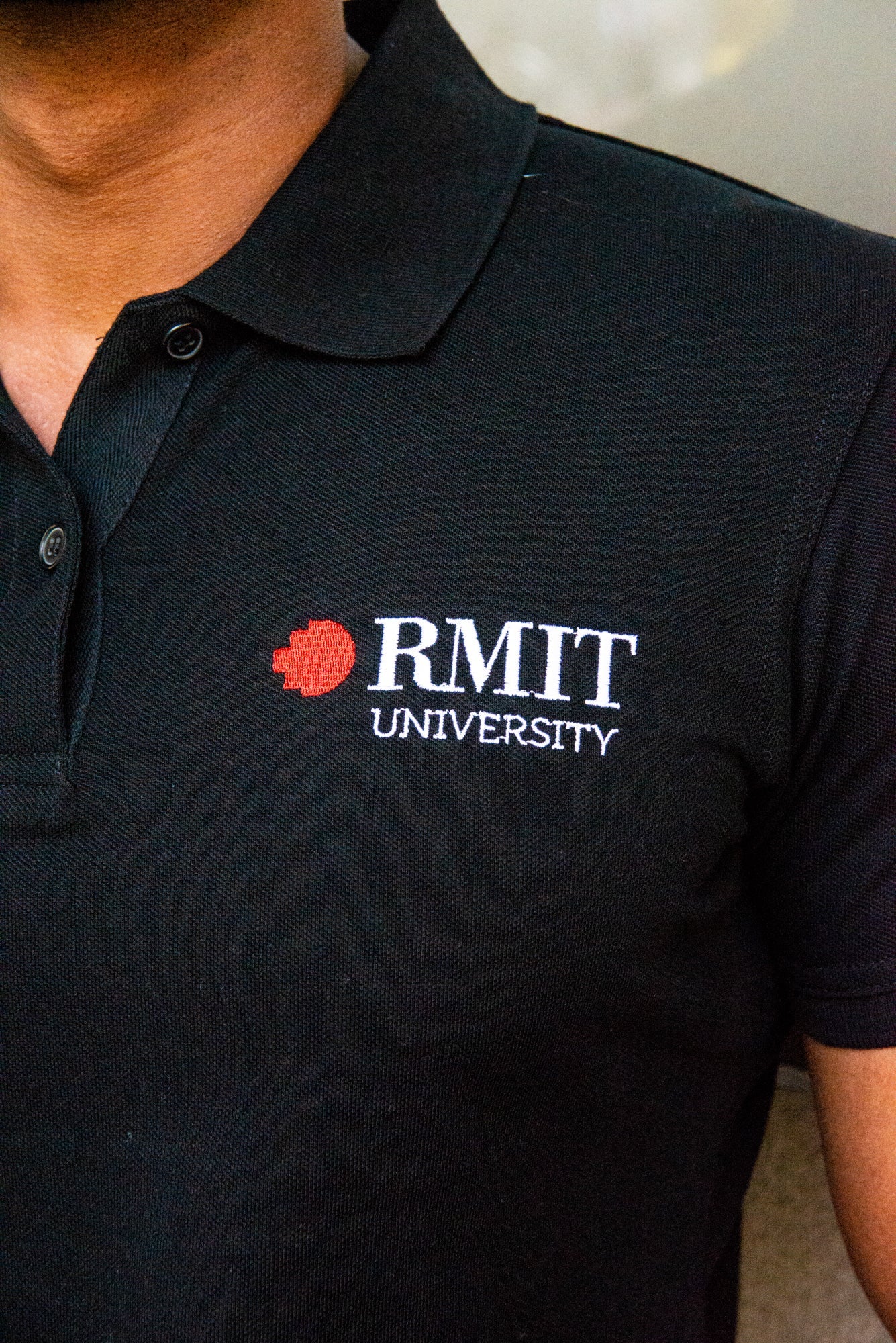 Detailed look at the RMIT University logo on the black polo, highlighting embroidery precision. The model's muscular build enhances the professional image associated with this uniform, distributed by the RMIT Store.