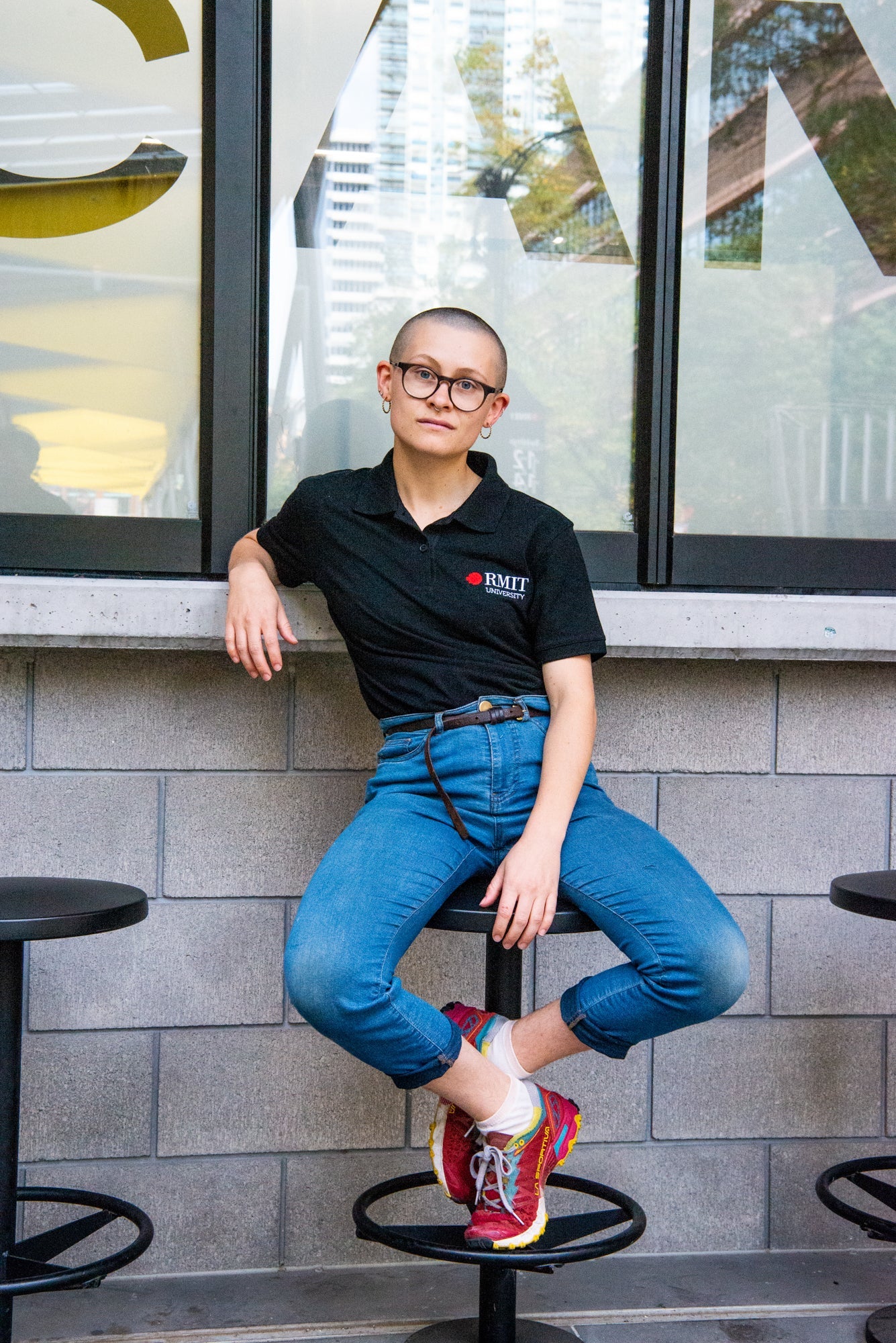 Model sitting on a stool wearing a black RMIT Polo with blue jeans and colourful sneakers. The ensemble reflects a relaxed and trendy style, with the polo available at the RMIT Store.