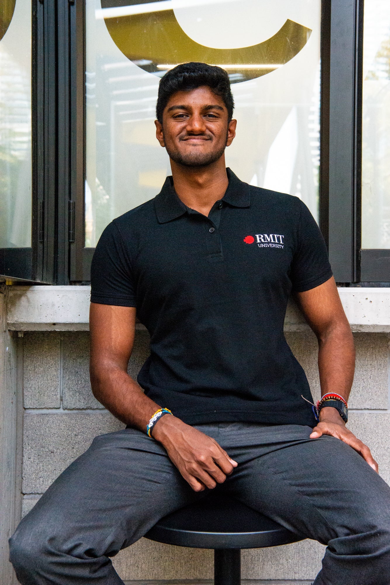 Model sitting straight in a black RMIT Polo adorned with the RMIT University logo, paired with grey trousers. The setting accentuates the uniform's professional appearance, available from the RMIT Store.