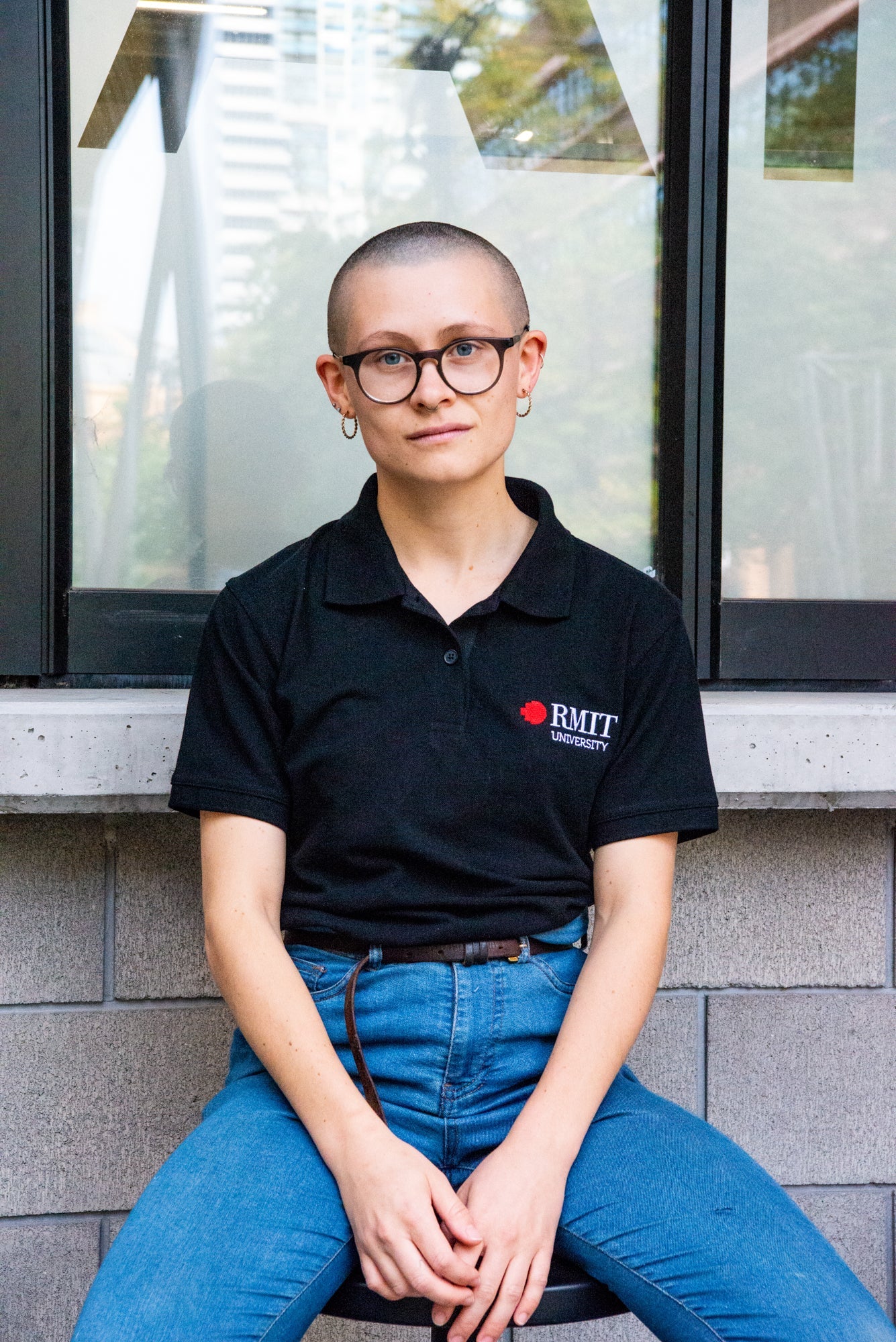 Full view of a model sitting in a black RMIT Polo with the RMIT University logo, paired with blue jeans. The setting provides a casual urban vibe, featuring this classic piece from the RMIT Store.