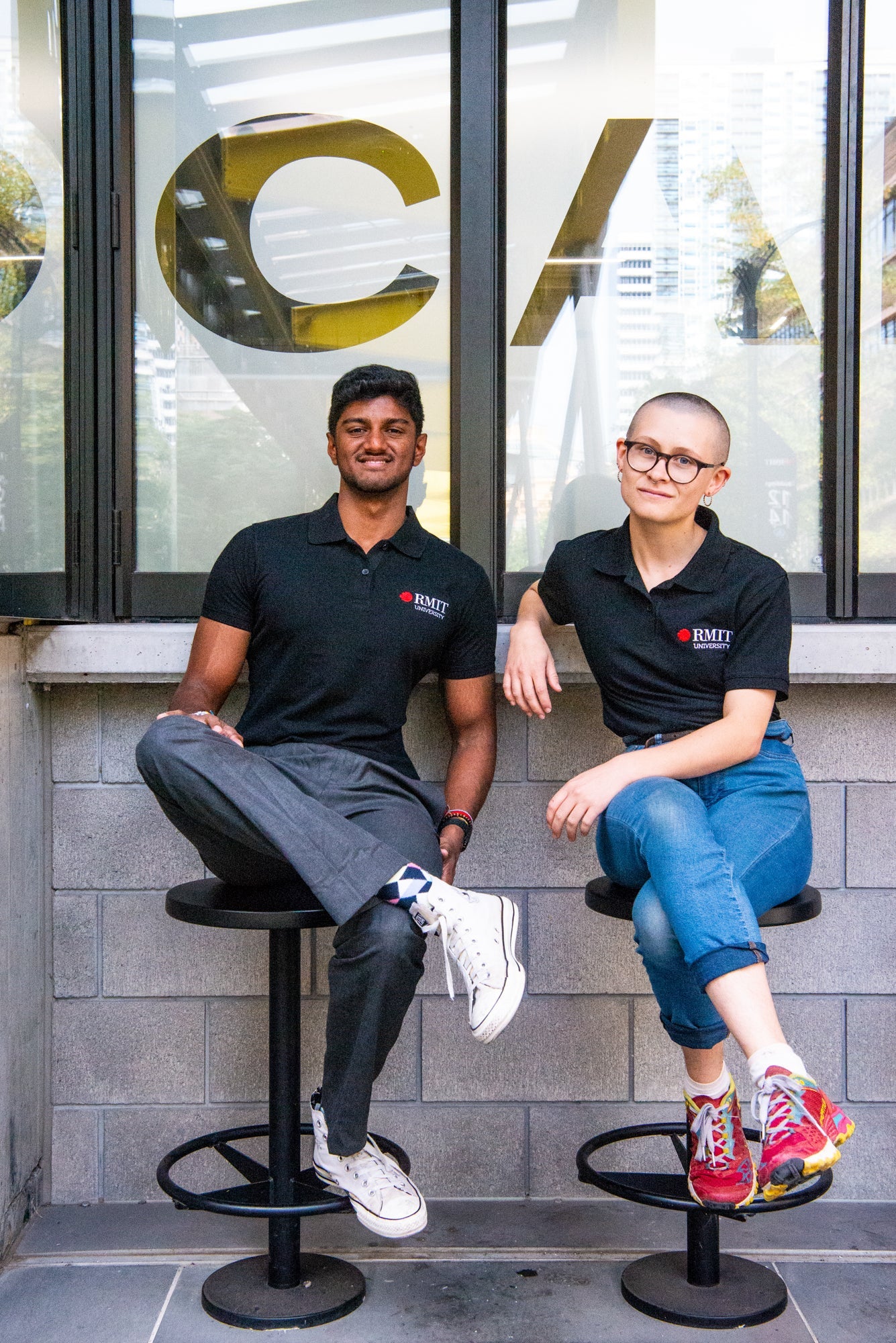 Two models seated side by side, both wearing black RMIT Polo shirts with the RMIT University logo. The image captures their casual posture and diverse styling, reflecting the polo's versatility as a uniform. Available at the RMIT Store.