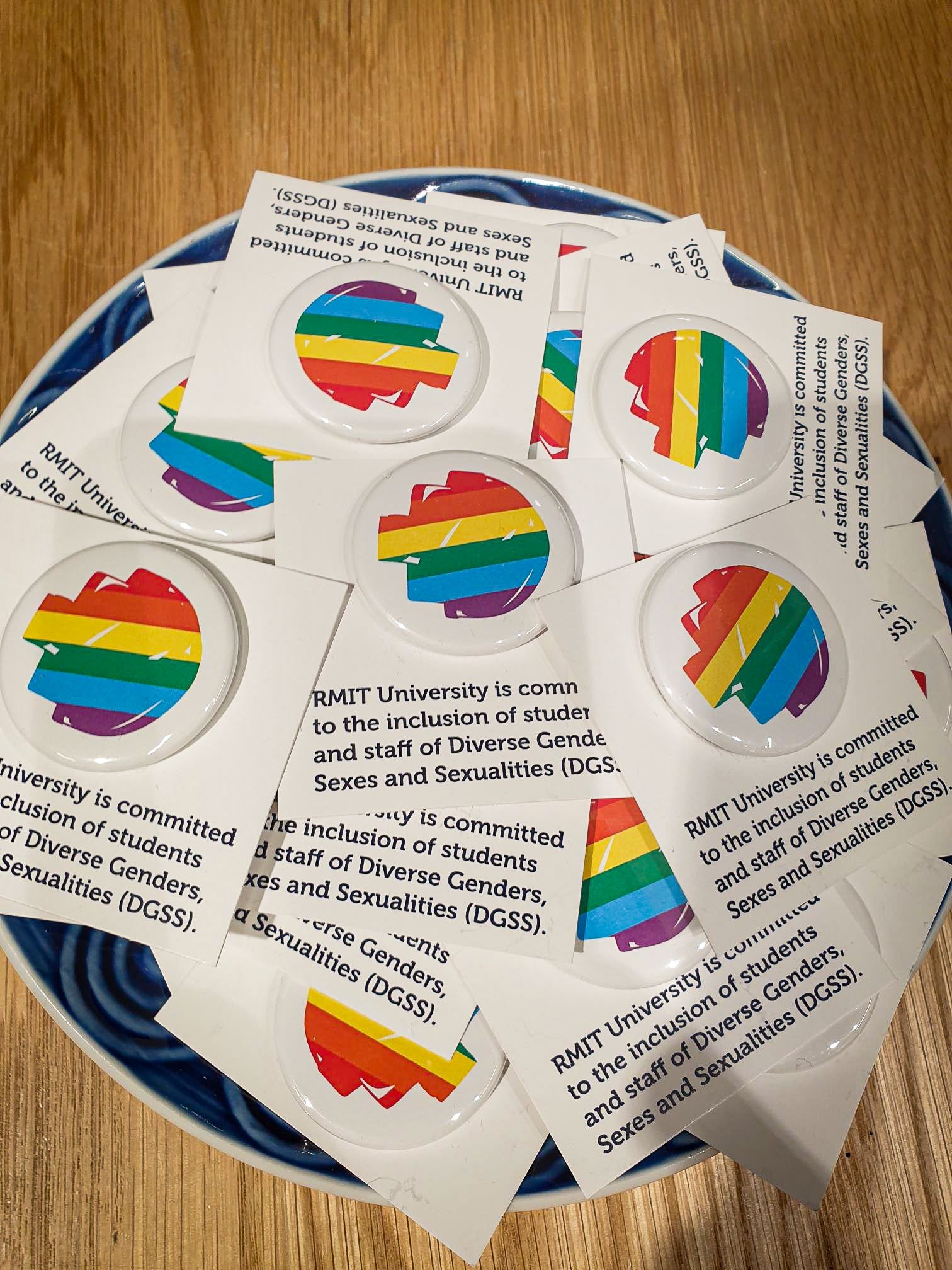 A collection of colourful pride badges featuring rainbow pixel designs scattered across a blue ceramic plate, placed on a wooden tabletop. Available from the RMIT Store.