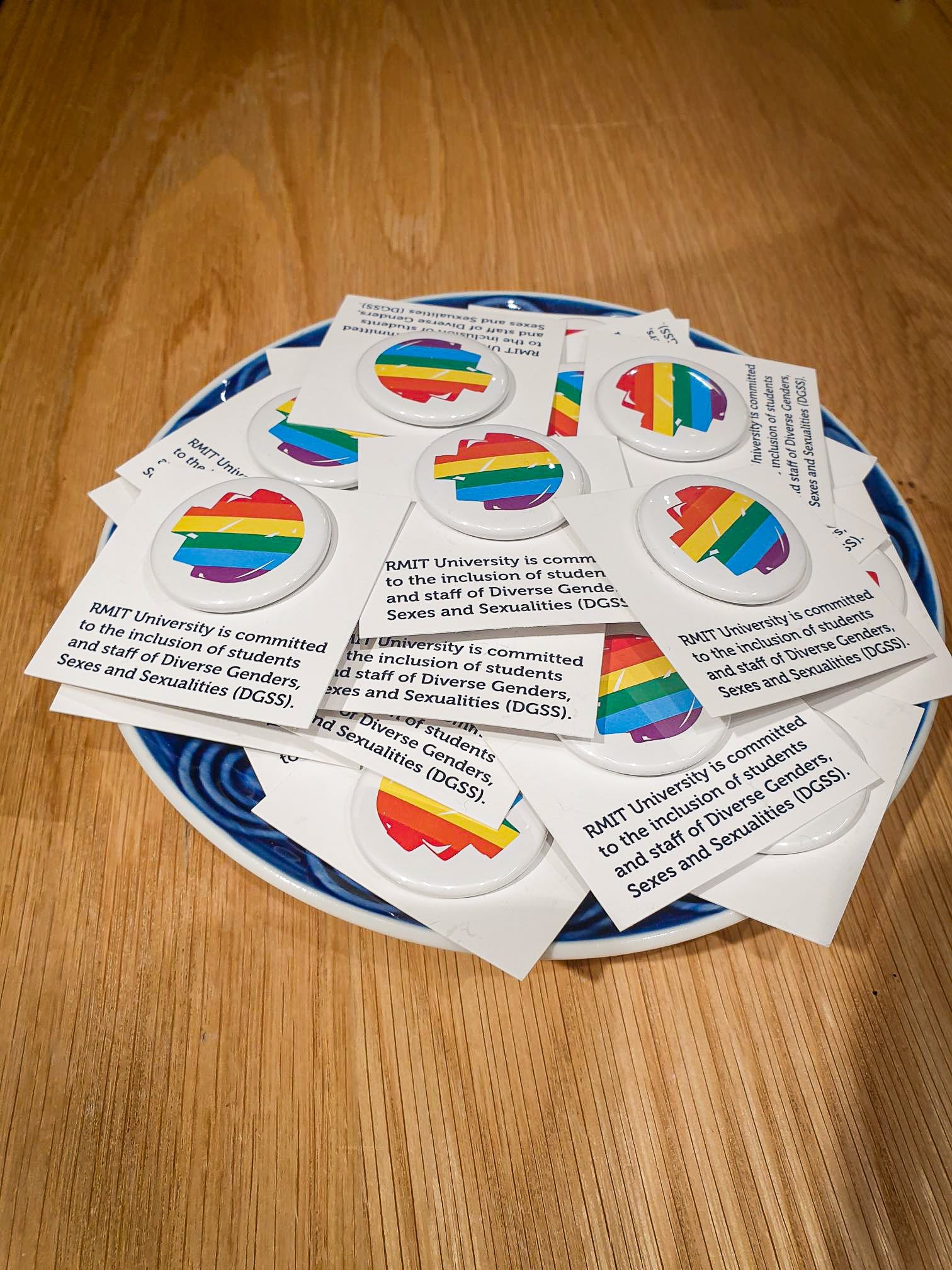 A collection of colourful pride badges featuring rainbow pixel designs scattered across a blue ceramic plate, placed on a wooden tabletop. Available from the RMIT Store.