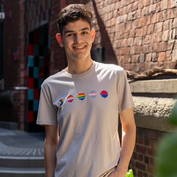 RMIT Store Pride Unity Tee in Bone, modelled by a person standing with one hand in his pocket, facing slightly sideways against a neutral background. The bone-coloured cotton t-shirt features five vibrant pride flags printed across the chest, representing different LGBTIQA+ identities. The tee has a relaxed fit, short sleeves, and a round neckline, offering both style and comfort. The model's pose, with a hand in his pocket, highlights the tee’s casual and inclusive design.
