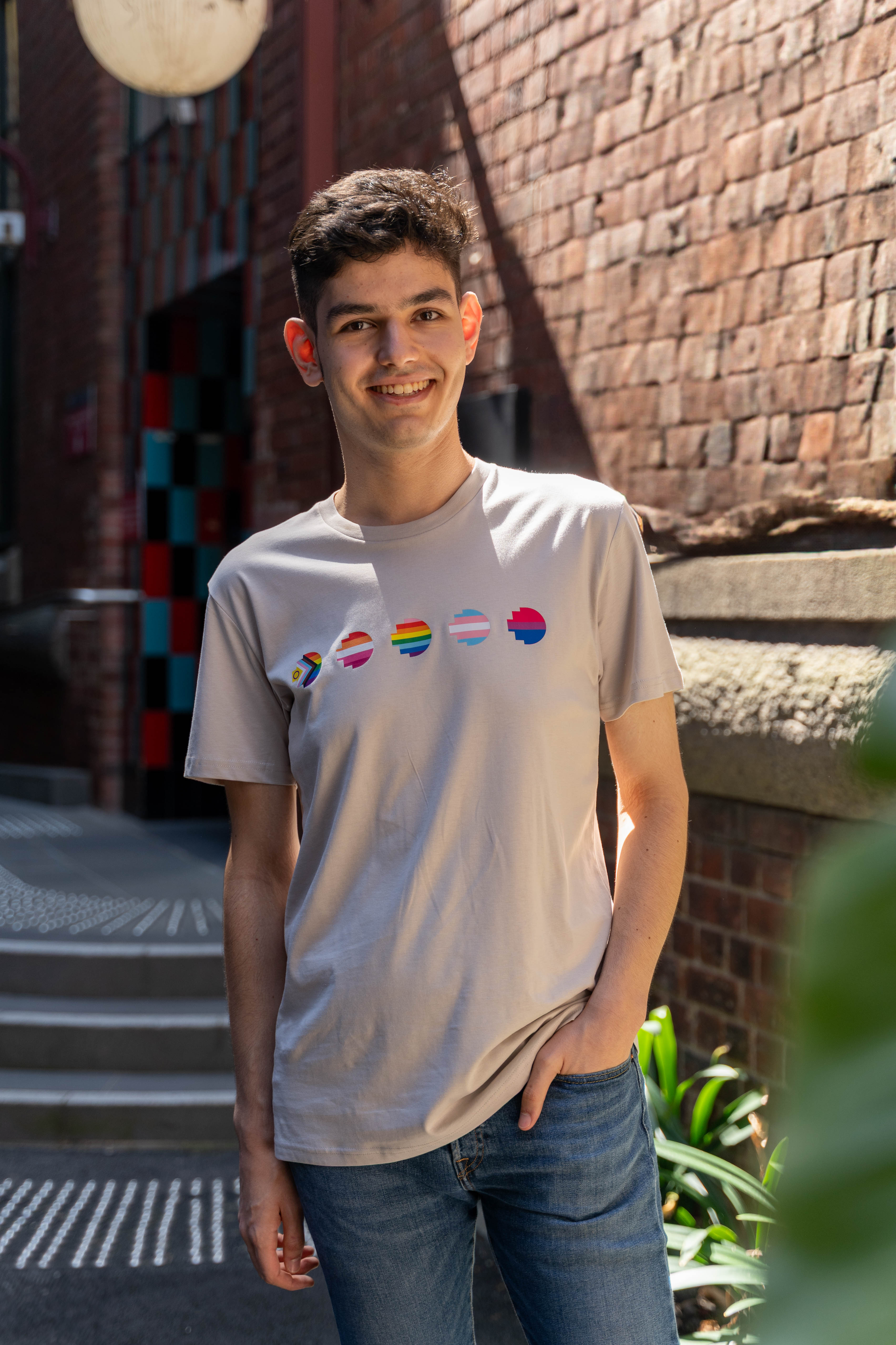RMIT Store Pride Unity Tee in Bone, modelled by a person standing with one hand in his pocket, facing slightly sideways against a neutral background. The bone-coloured cotton t-shirt features five vibrant pride flags printed across the chest, representing different LGBTIQA+ identities. The tee has a relaxed fit, short sleeves, and a round neckline, offering both style and comfort. The model's pose, with a hand in his pocket, highlights the tee’s casual and inclusive design.