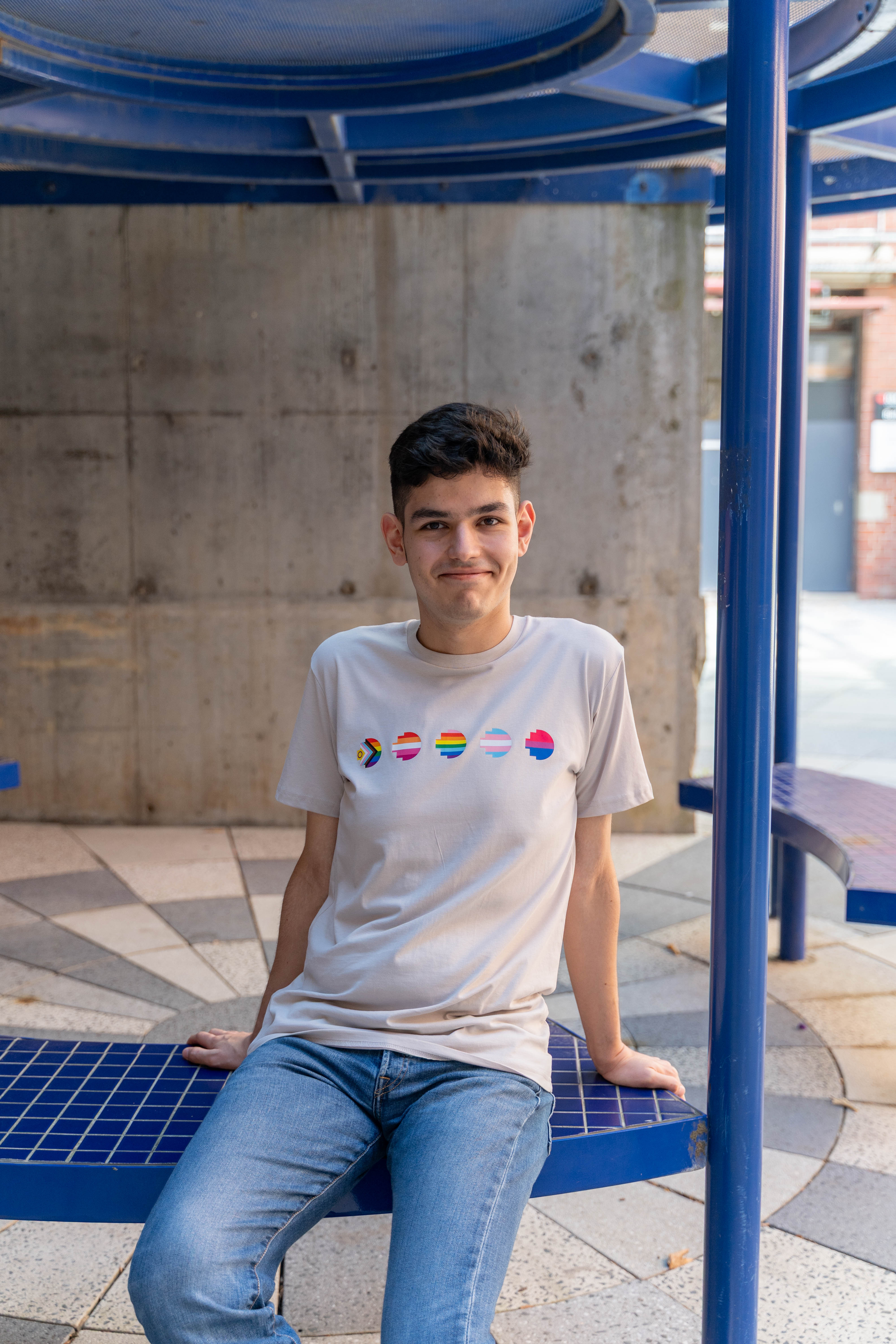 RMIT Store Pride Unity Tee in Bone, modelled by a person sitting and smiling against a neutral backdrop. The bone-coloured cotton t-shirt features five vibrant pride flags printed across the chest, representing different LGBTIQA+ identities. The tee has a relaxed fit, round neckline, and short sleeves, combining comfort and style. The model's seated, smiling pose and the wider shot highlight the tee's inclusivity and unity message.