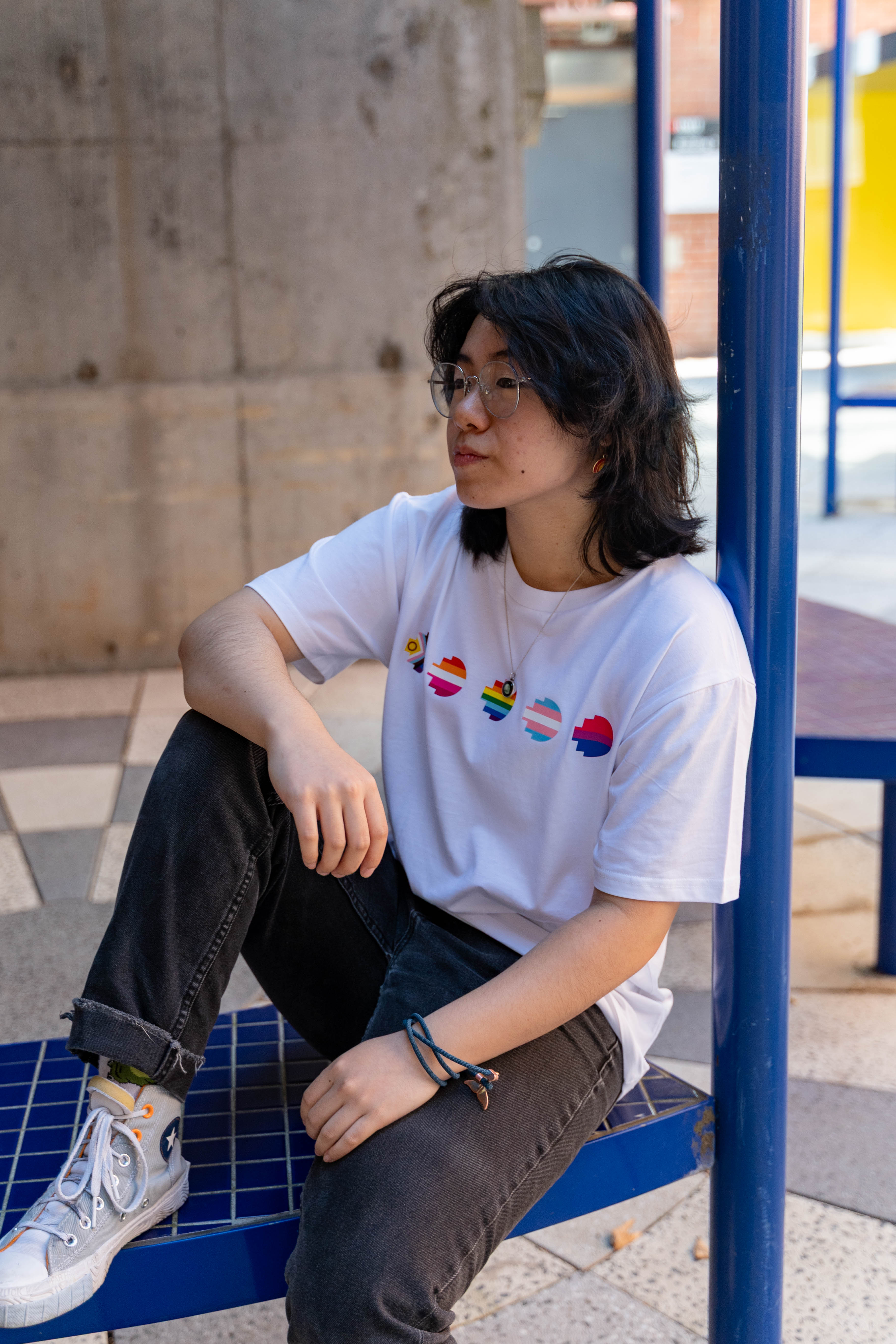 Model wears the Pride Unity Tee in limited edition white, featuring colourful pride emblem prints on the chest while seated on a blue mesh bench. Available at the RMIT Store.