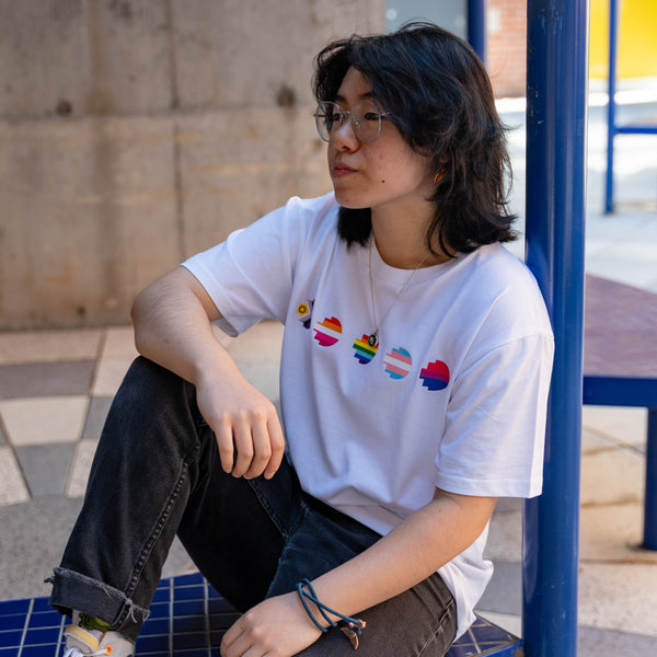 Model wears the Pride Unity Tee in limited edition white, featuring colourful pride emblem prints on the chest while seated on a blue mesh bench. Available at the RMIT Store.