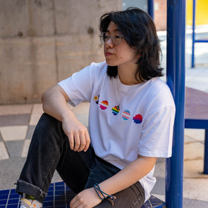 Model wears the Pride Unity Tee in limited edition white, featuring colourful pride emblem prints on the chest while seated on a blue mesh bench. Available at the RMIT Store.
