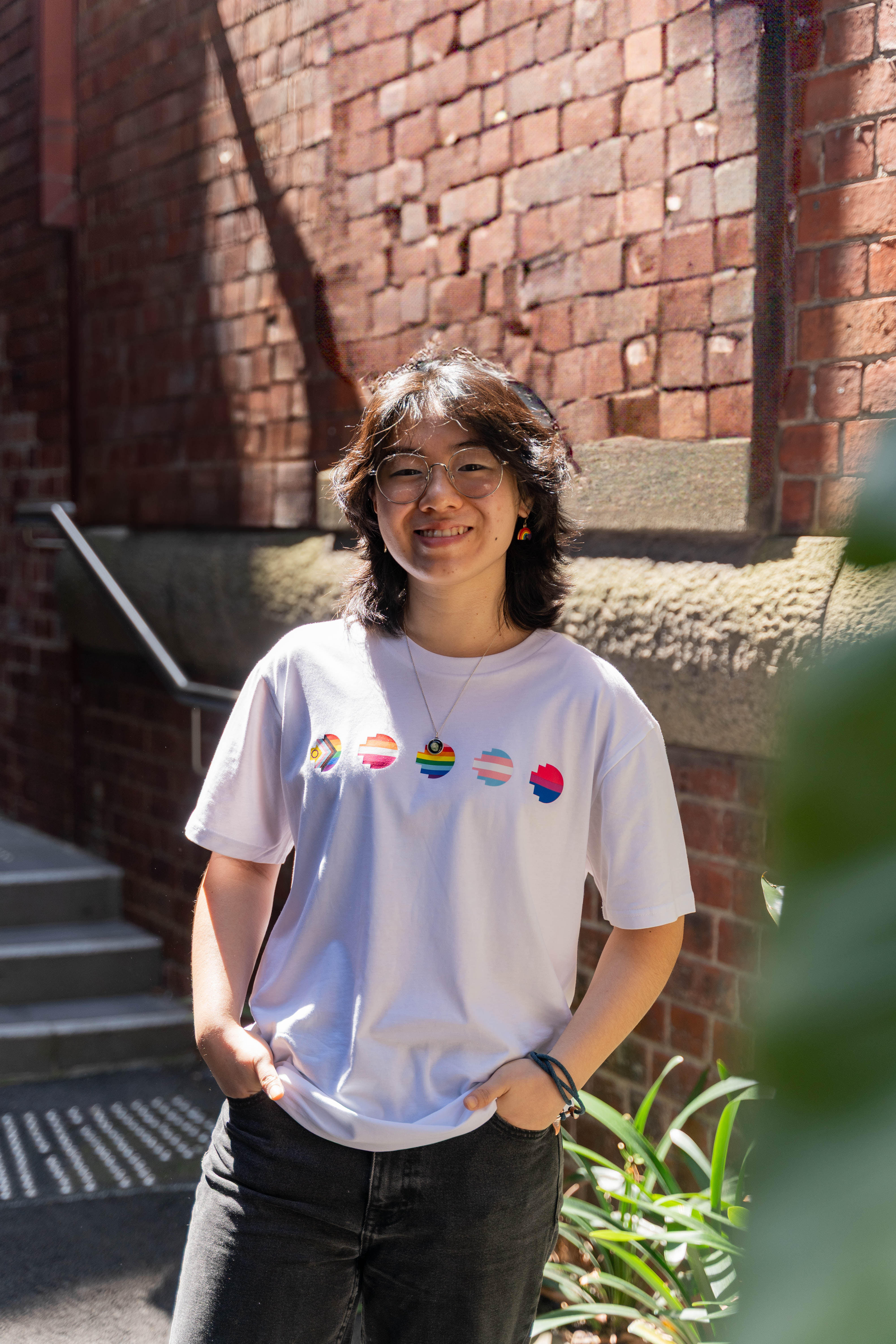 Model wears the Limited Edition Pride Unity Tee in white, showcasing colourful emblem designs on the front, standing in sunlight against a textured brick wall.