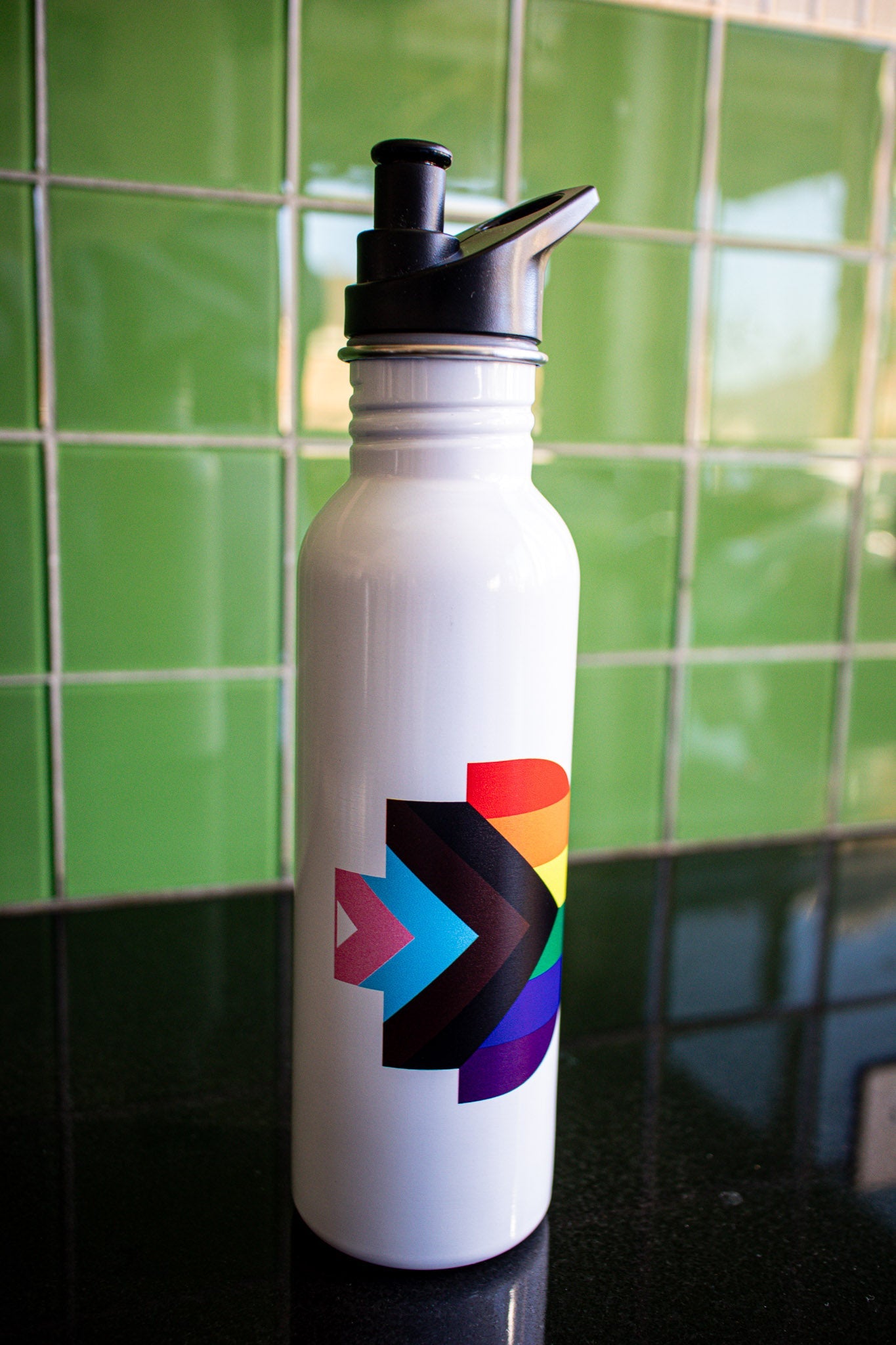 Front view of the Progress Pride bottle featuring the Progress Pride flag symbol printed on its white body. The bottle is placed on a black tabletop with a green background. The black cap adds contrast to the clean design. Available at the RMIT Store.