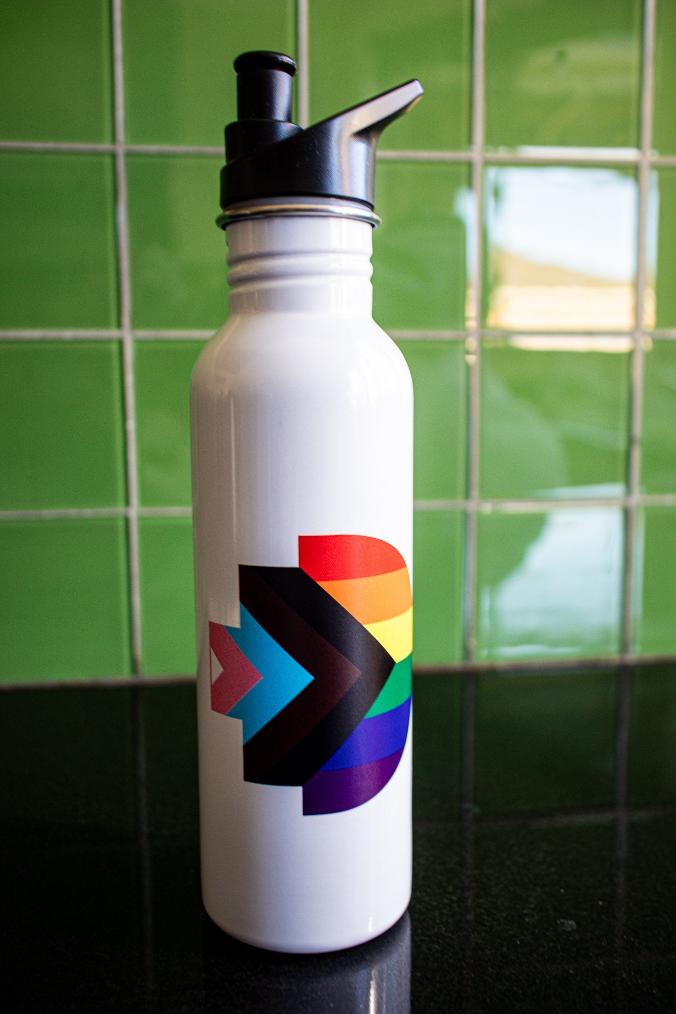 Front view of the Progress Pride bottle featuring the Progress Pride flag symbol printed on its white body. The bottle is placed on a black tabletop with a green background. The black cap adds contrast to the clean design. Available at the RMIT Store.
