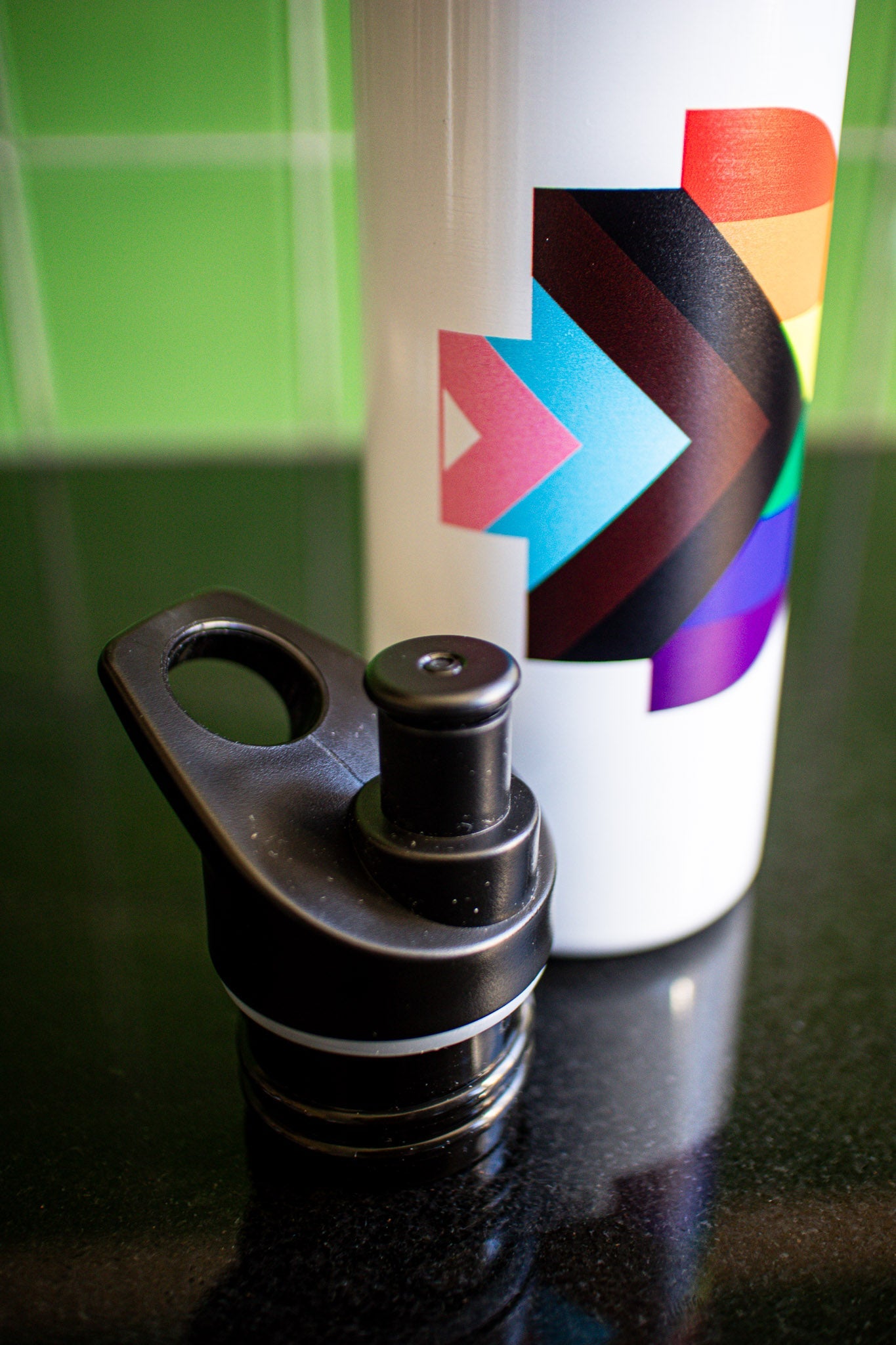Angled side view of the Progress Pride bottle showing the full Progress Pride logo on its white body, with the detached black cap placed to the left of the bottle. The scene is set on a black tabletop against a green background. Available at the RMIT Store.
