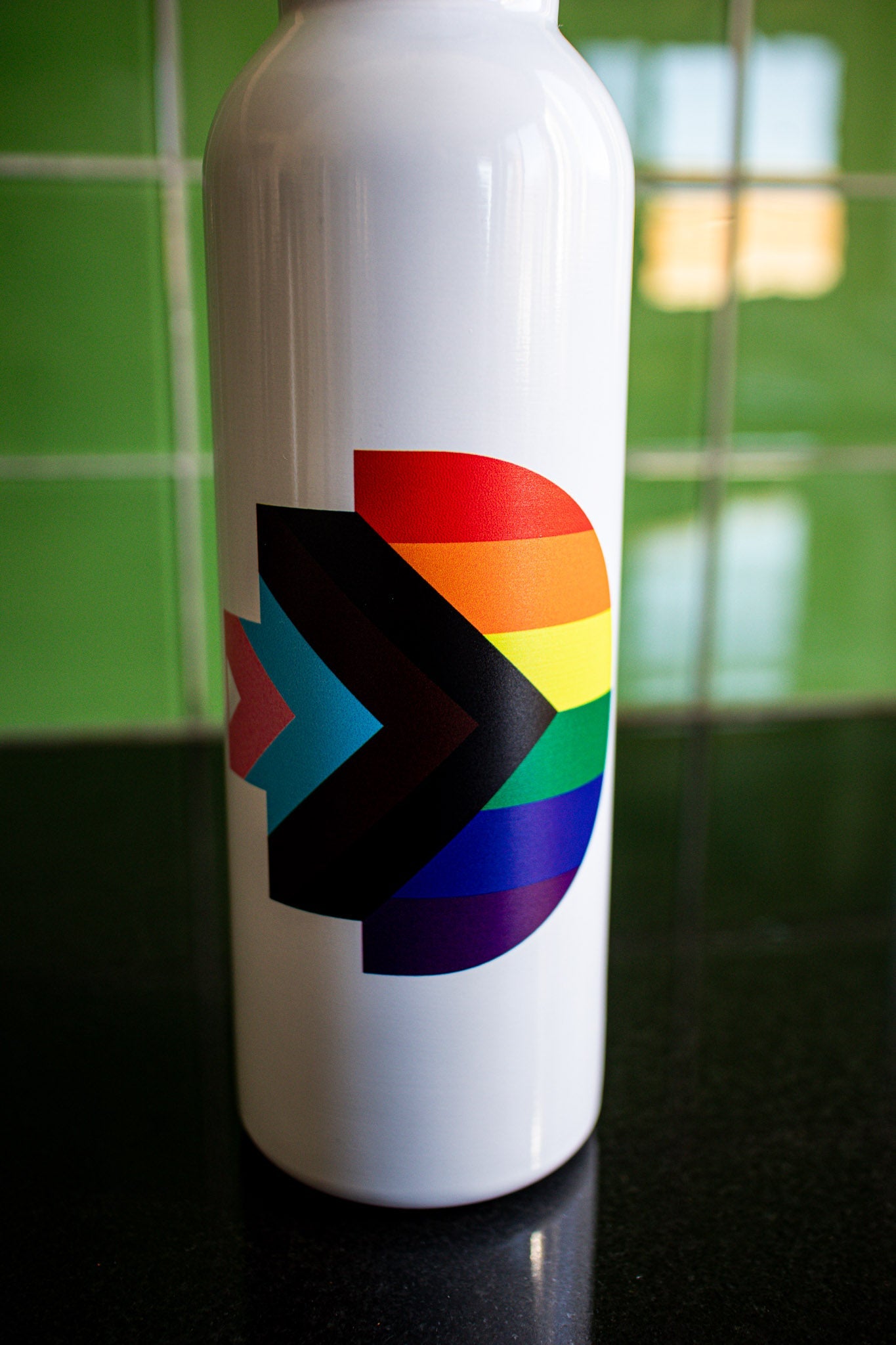 Close-up view of the Progress Pride symbol printed on the matte white body of the Progress Pride bottle. The detailed rainbow and chevron design is clearly visible, with the bottle positioned on a black tabletop against a green background. Available at the RMIT Store.