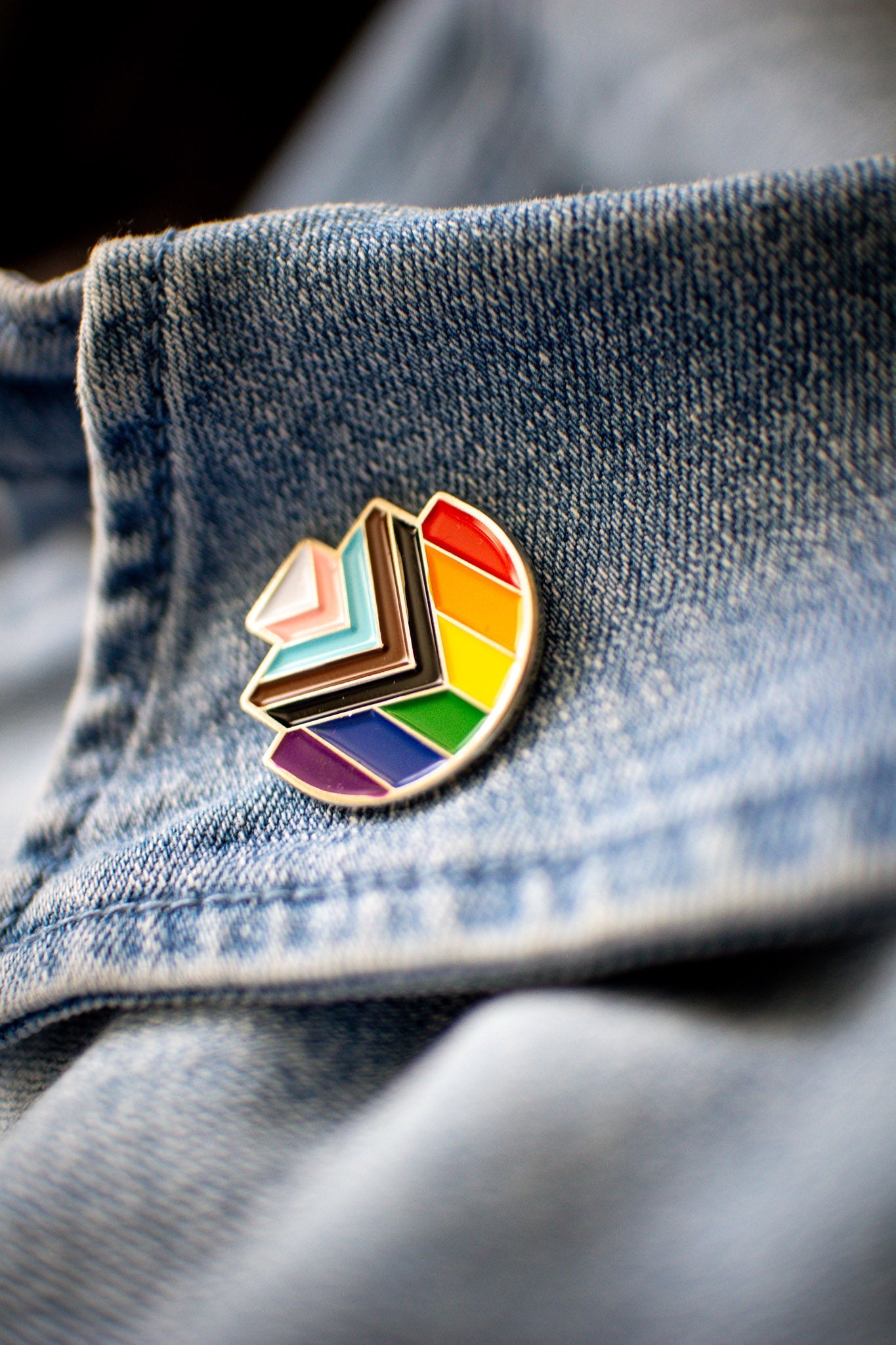 Progress Pride enamel pin in the shape of RMIT Pixel logo, featuring the rainbow flag with inclusive chevron design in black, brown, light blue, pink, and white, resting on a blue denim collar with visible stitching. Available at the RMIT Store.