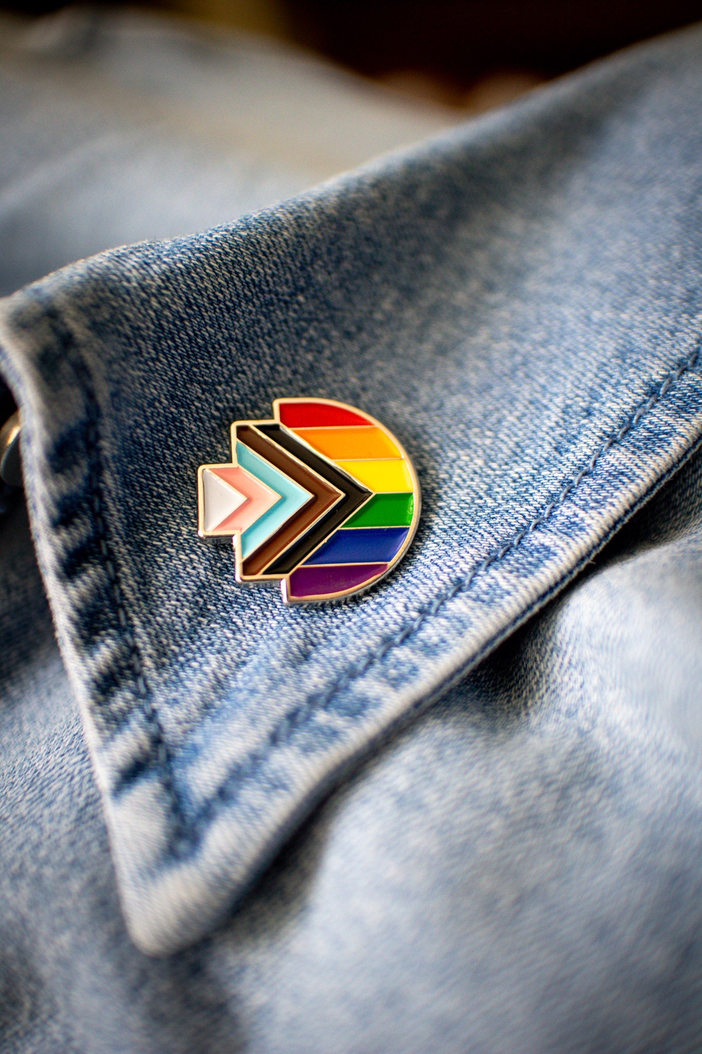 Progress Pride enamel pin in the shape of RMIT Pixel logo, featuring the rainbow flag with inclusive chevron design in black, brown, light blue, pink, and white, resting on a blue denim collar with visible stitching. Available at the RMIT Store.
