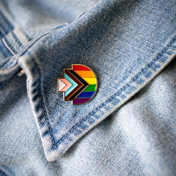 Progress Pride enamel pin in the shape of RMIT Pixel logo, featuring the rainbow flag with inclusive chevron design in black, brown, light blue, pink, and white, resting on a blue denim collar with visible stitching. Available at the RMIT Store.