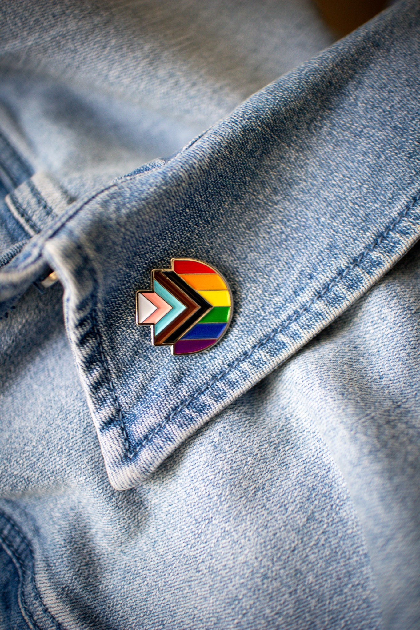 Progress Pride enamel pin in the shape of RMIT Pixel logo, featuring the rainbow flag with inclusive chevron design in black, brown, light blue, pink, and white, resting on a blue denim collar with visible stitching. Available at the RMIT Store.