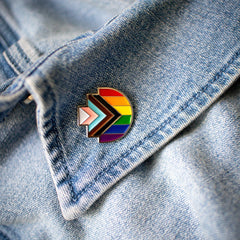 Progress Pride enamel pin in the shape of RMIT Pixel logo, featuring the rainbow flag with inclusive chevron design in black, brown, light blue, pink, and white, resting on a blue denim collar with visible stitching. Available at the RMIT Store.