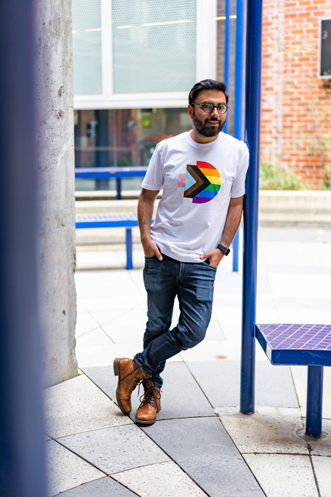 Full view of a model wearing the Progress Pride Tee, leaning casually against a blue pillar in an urban setting. The white tee displays a large, centrally printed rainbow-coloured RMIT pixel logo. Available at RMIT Store.