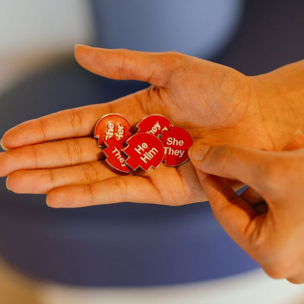 Close-up of a hand holding a collection of RMIT Store’s pronoun pins, including red pixelated designs for “she/her,” “he/him,” “they/them", "she/they" and "he/they". Each pin features white printed text on a red pixelated background.