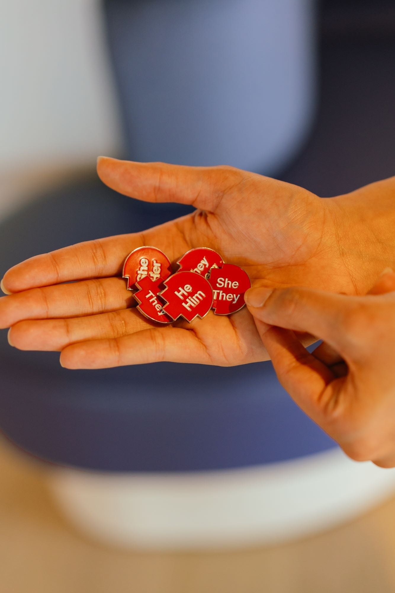 Close-up of a hand holding a collection of RMIT Store’s pronoun pins, including red pixelated designs for “she/her,” “he/him,” “they/them", "she/they" and "he/they". Each pin features white printed text on a red pixelated background.