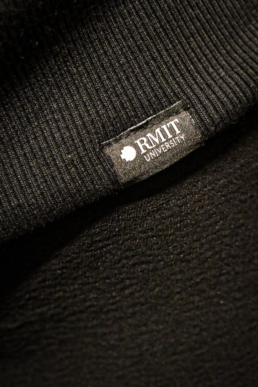 Close-up view of the Puff Pixel Hoodie in black, showing the small white embroidered RMIT logo near the bottom hem. Available from the RMIT Store.