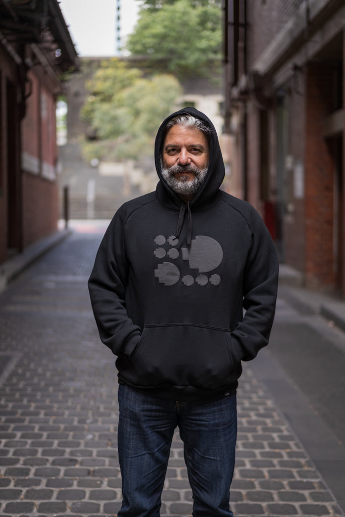 Model wearing the Puff Pixel Hoodie in black with the hood up, smiling while standing in an outdoor campus corridor. Available at RMIT Store.
